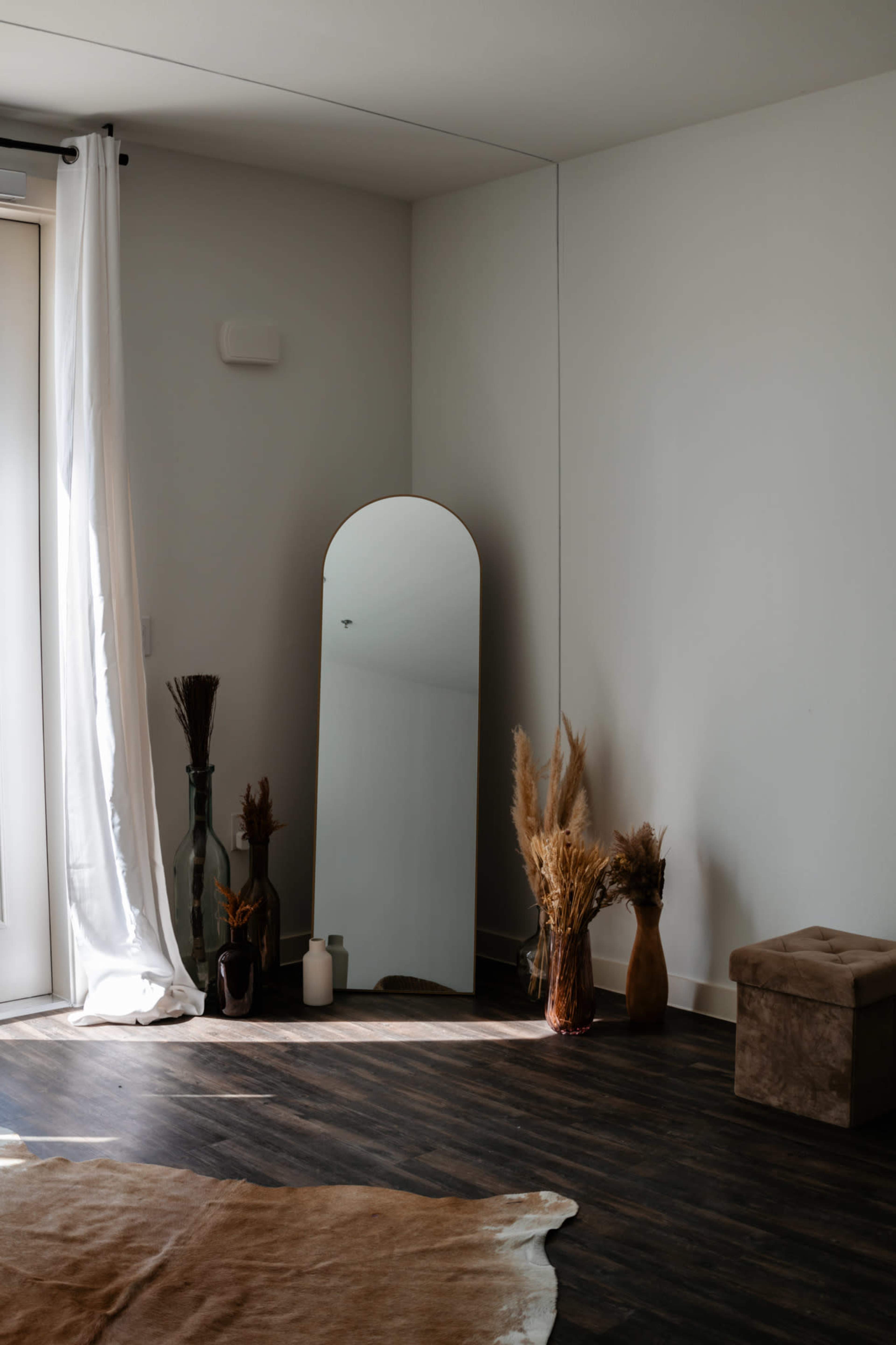 A large, arched mirror stands against a wall in a spacious room, surrounded by decorative plants and a small stool.