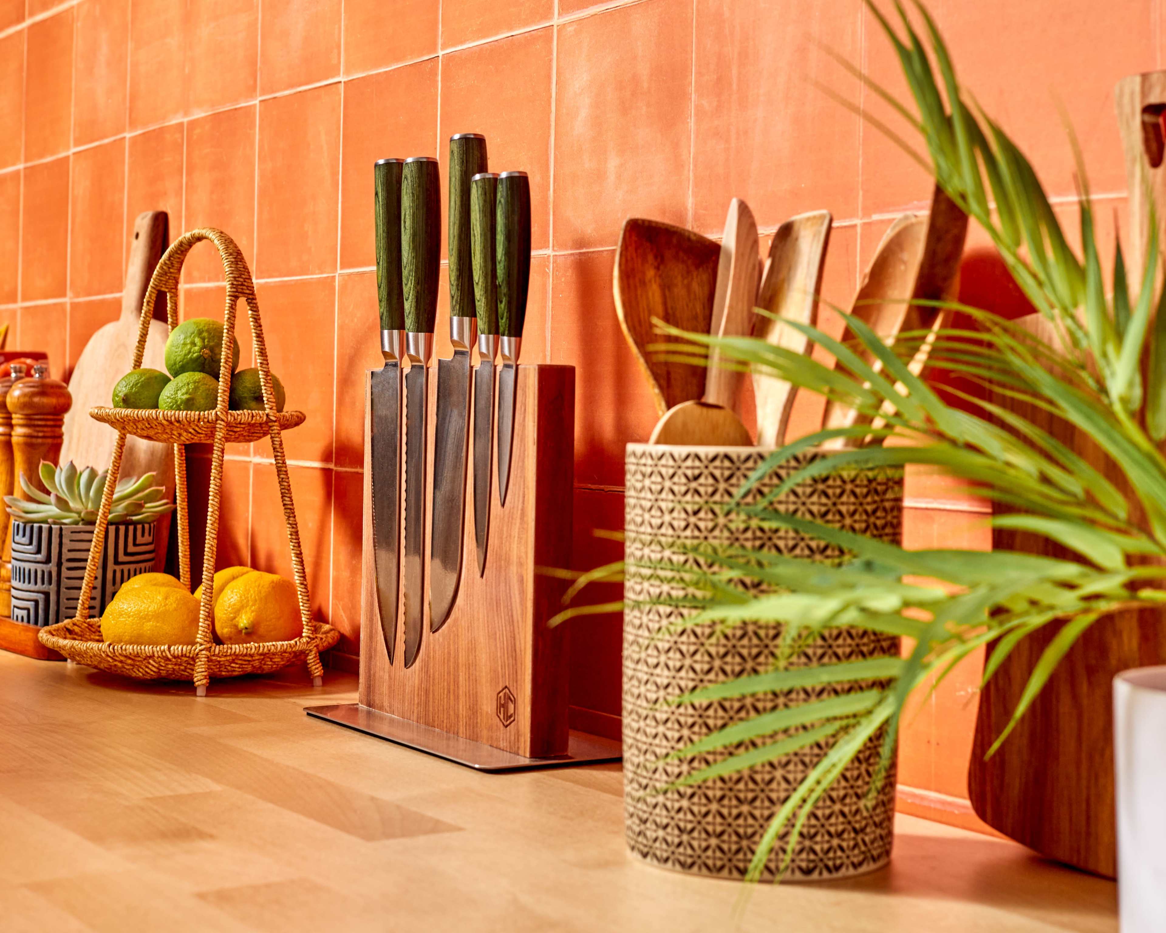 A kitchen countertop featuring a knife block with five knives, a bamboo utensil holder, and a two-tier fruit basket containing lemons and limes against a tiled orange backdrop.