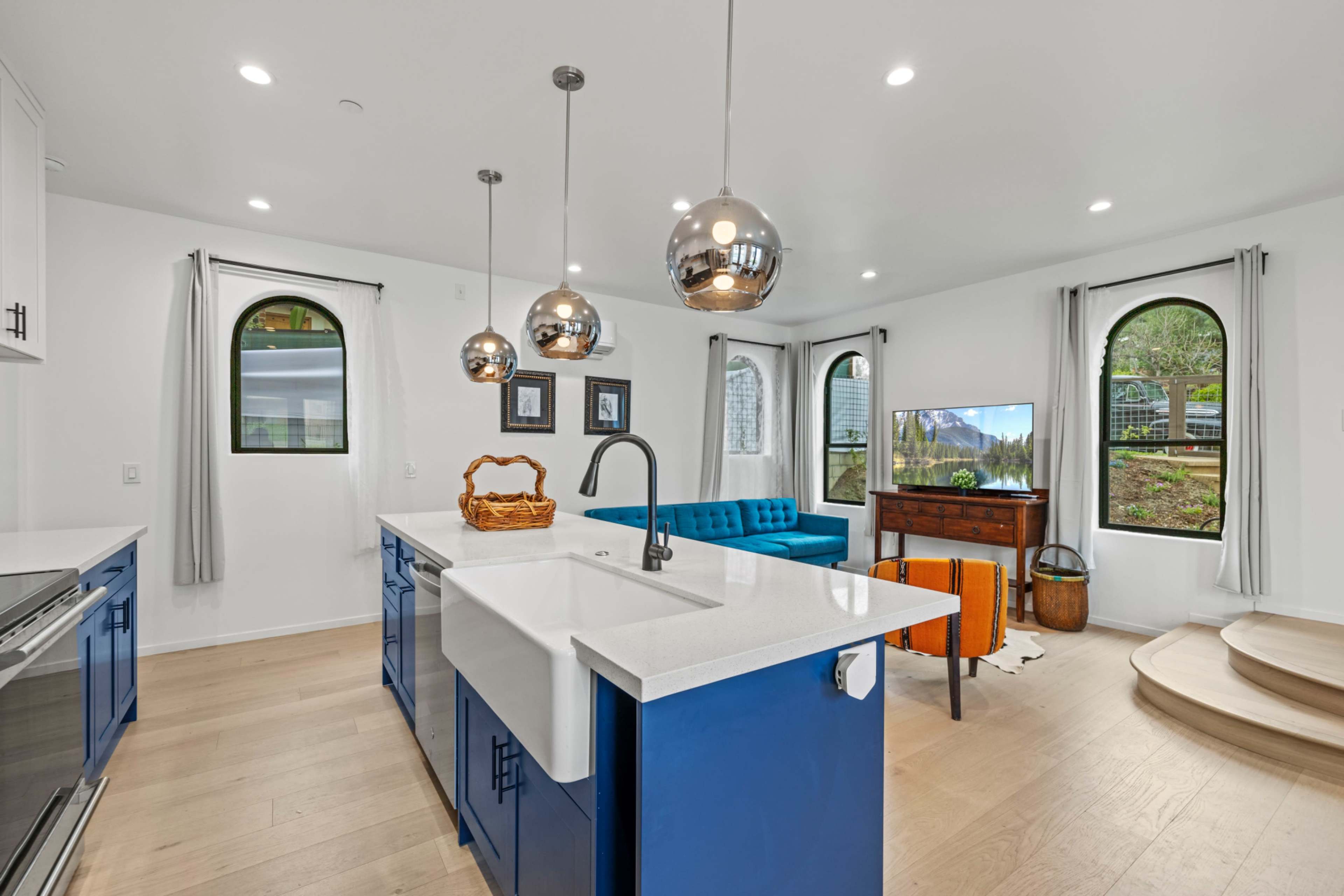 The image shows a modern kitchen and living area featuring blue cabinetry, a large farmhouse sink, metallic pendant lights, and a comfortable seating area with a television.