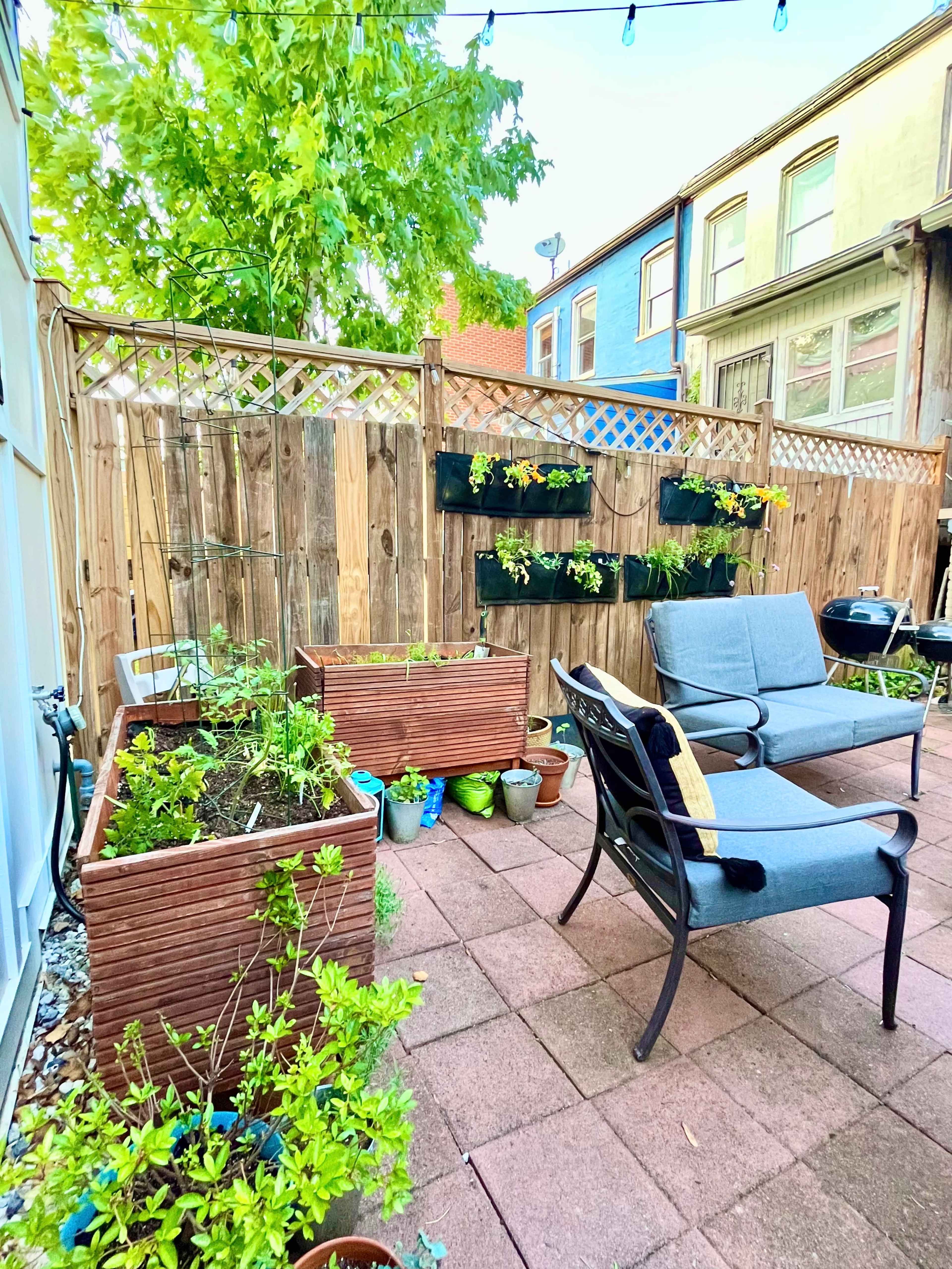 A small garden area with raised planting boxes, a seating arrangement of two chairs, and a wooden fence adorned with hanging planters.