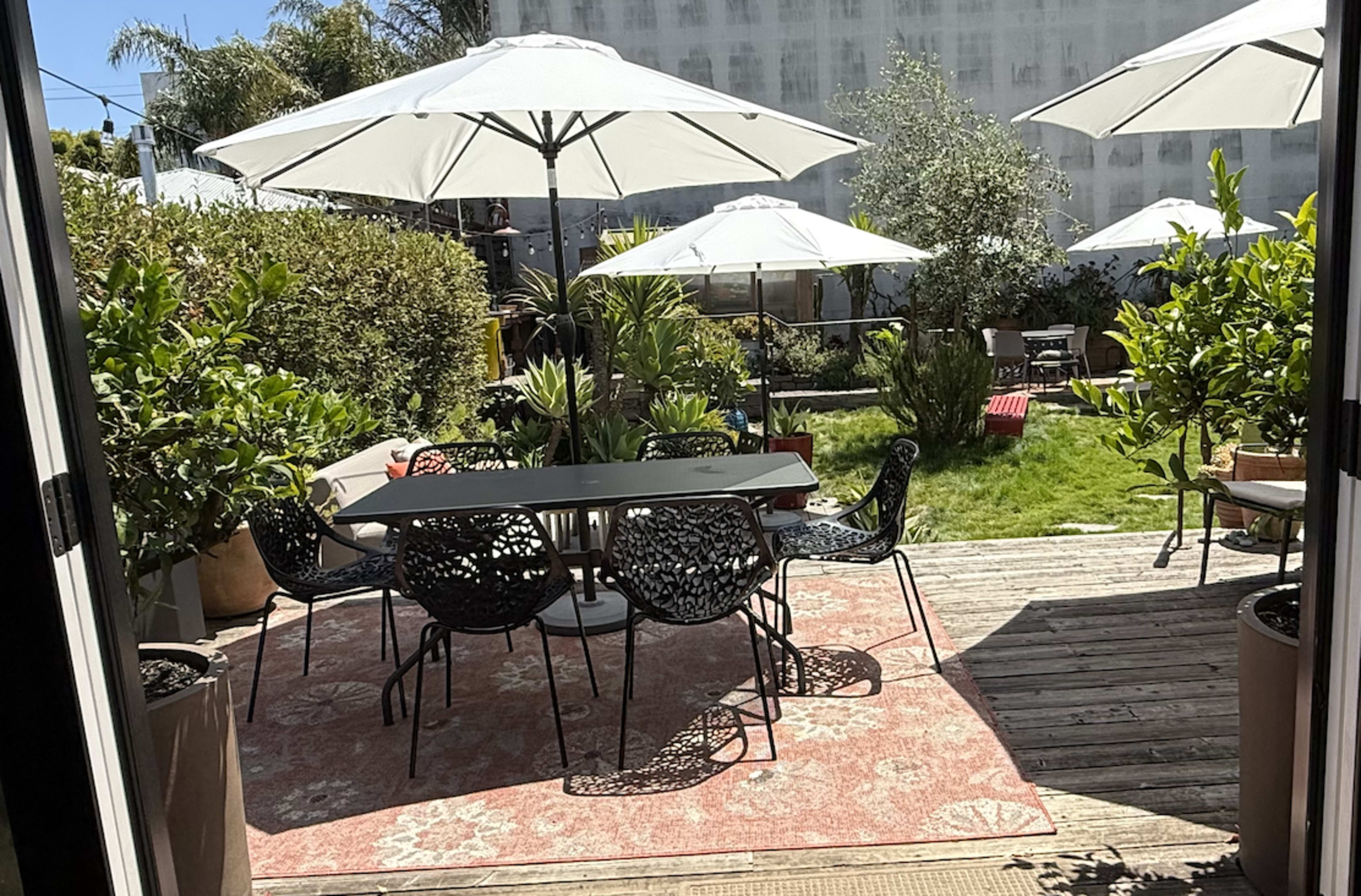 A patio area is visible, featuring a large dining table surrounded by black chairs, sun umbrellas, and various plants in pots.