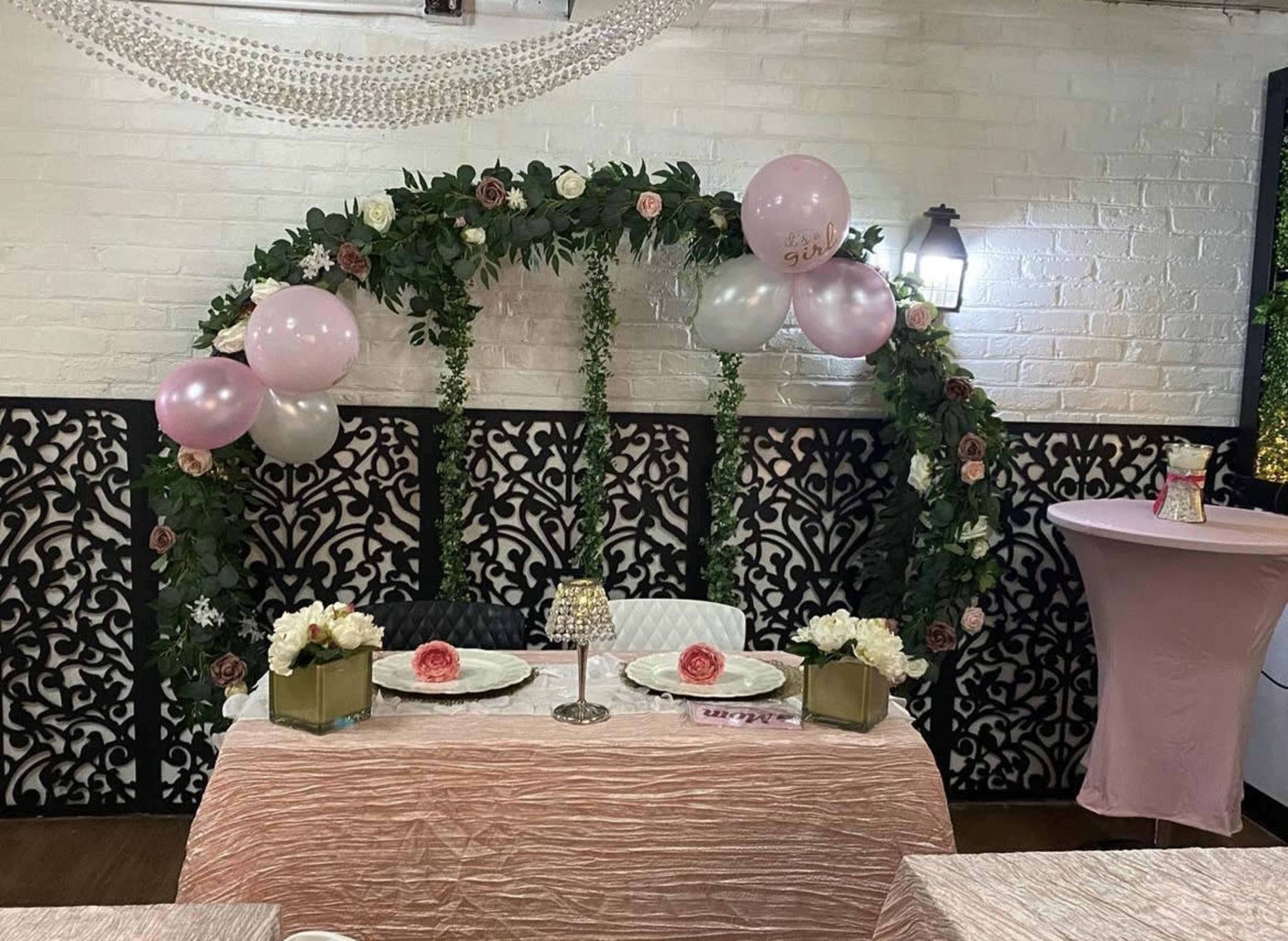A decorated table with floral arrangements and balloons is set under an arch of greenery in a banquet area.