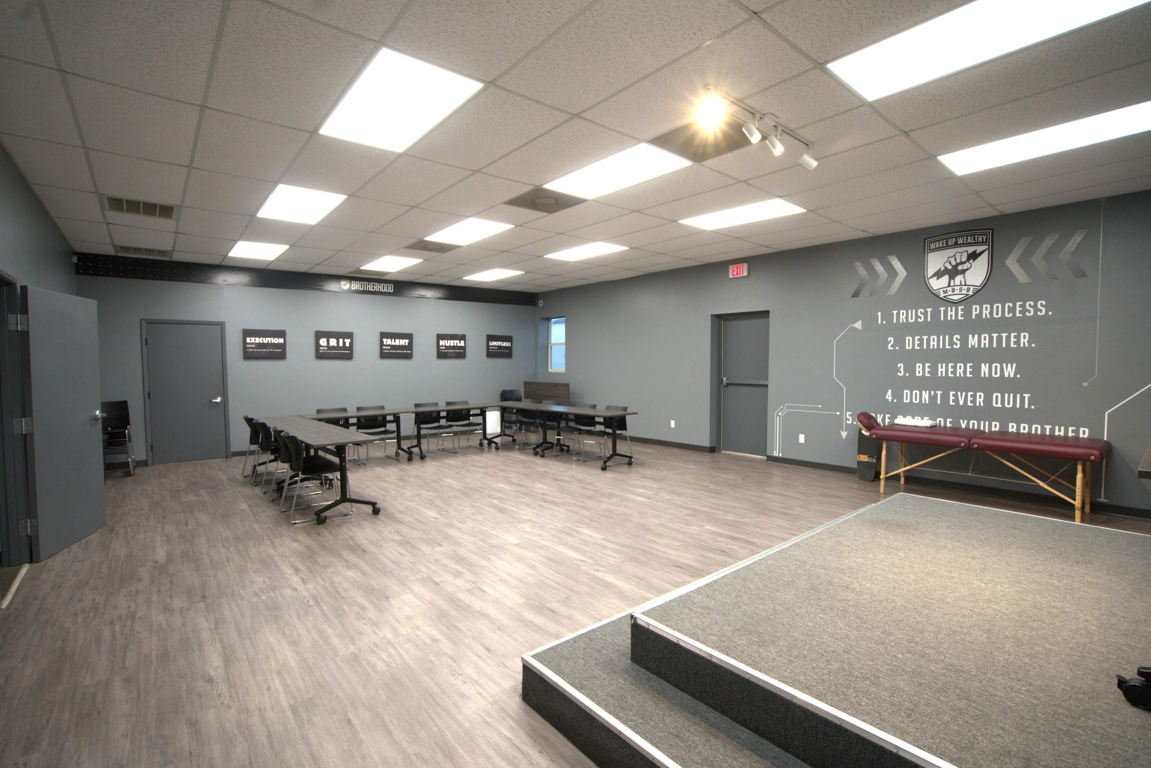 The image shows a spacious training room with gray walls, a long table surrounded by chairs, and motivational quotes displayed on the walls.