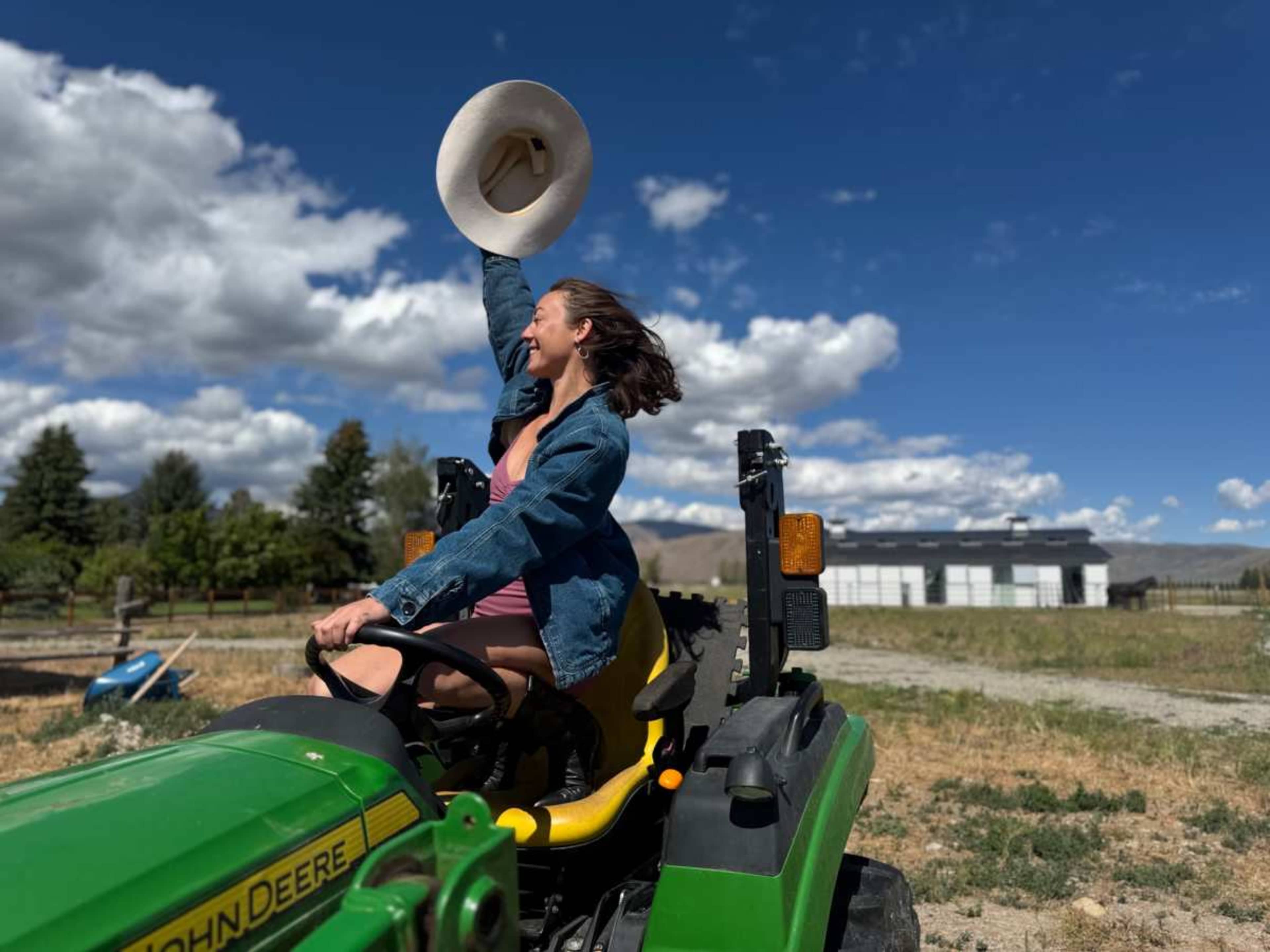 A person is driving a green tractor while holding a large hat in one hand against a backdrop of blue skies and scattered clouds.