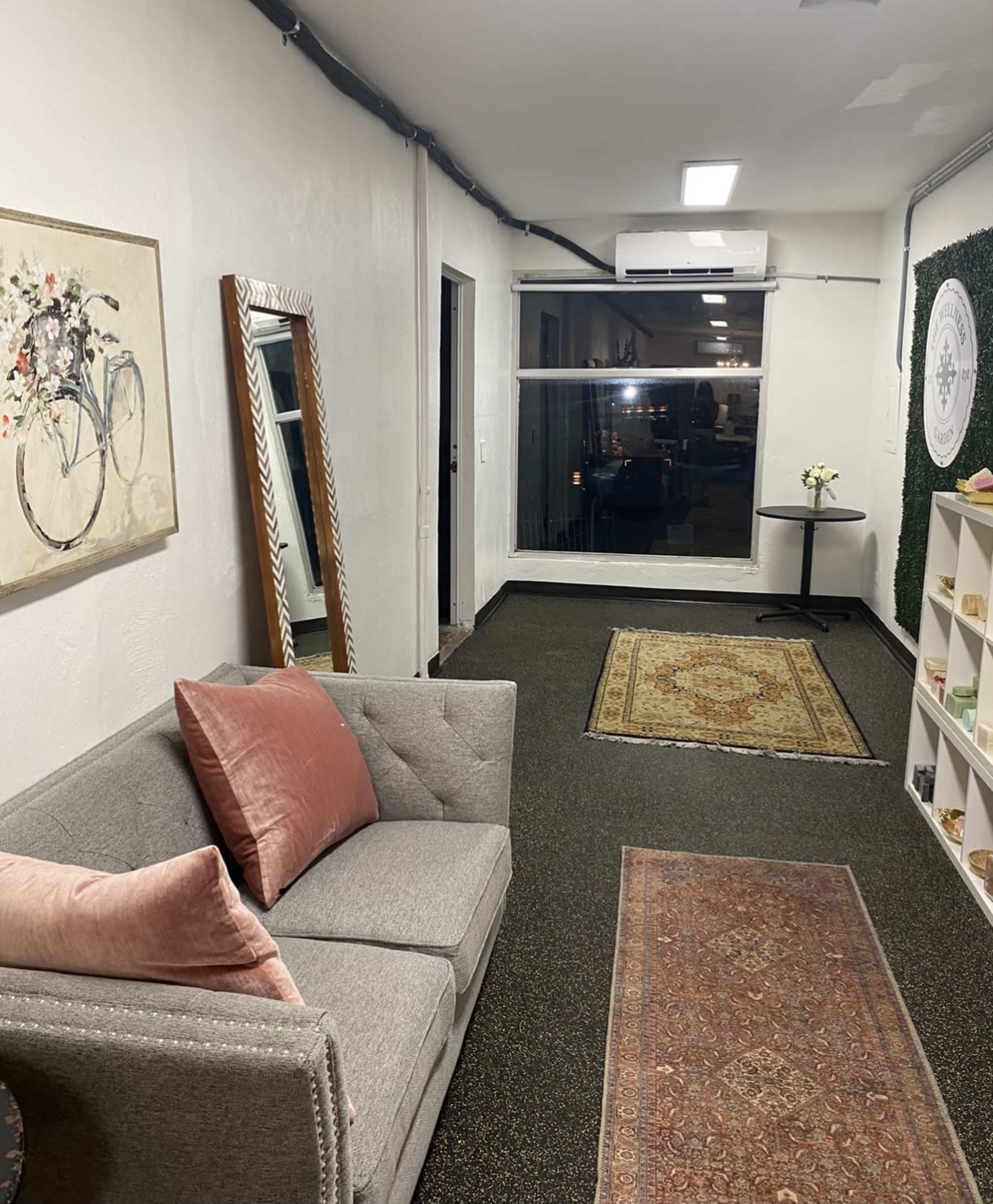 The image shows a small, well-lit room featuring a gray couch, a decorative mirror, a window with sheer curtains, and a small table with a vase of flowers.