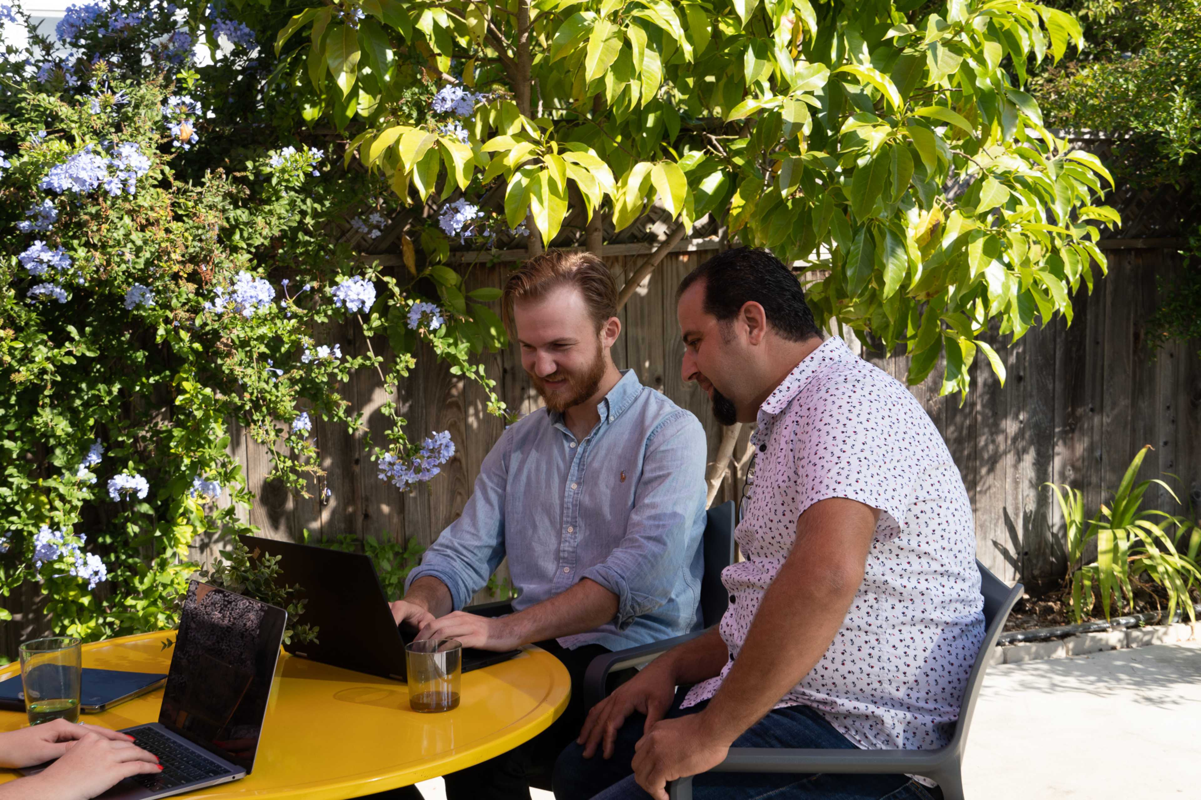 Two men are seated at a yellow table outdoors, working on laptops under a tree with blue flowers.