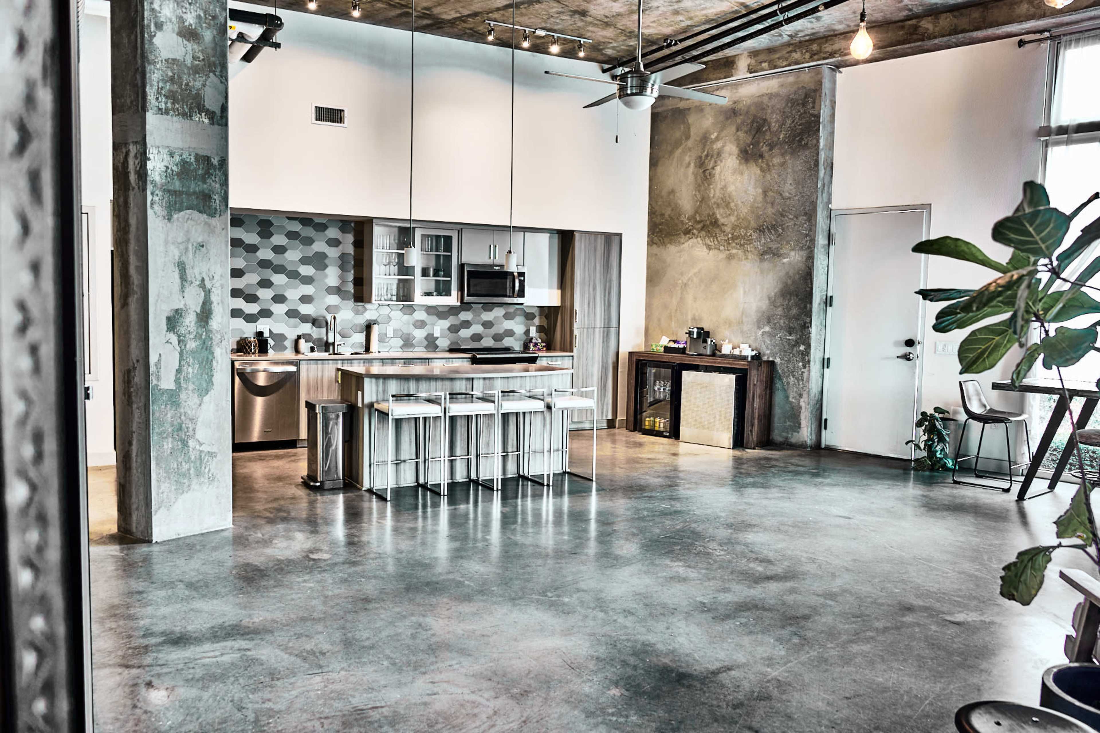 The image shows a modern kitchen and dining area with concrete floors, pendant lighting, a central island with bar stools, and a mix of textured walls and cabinetry.