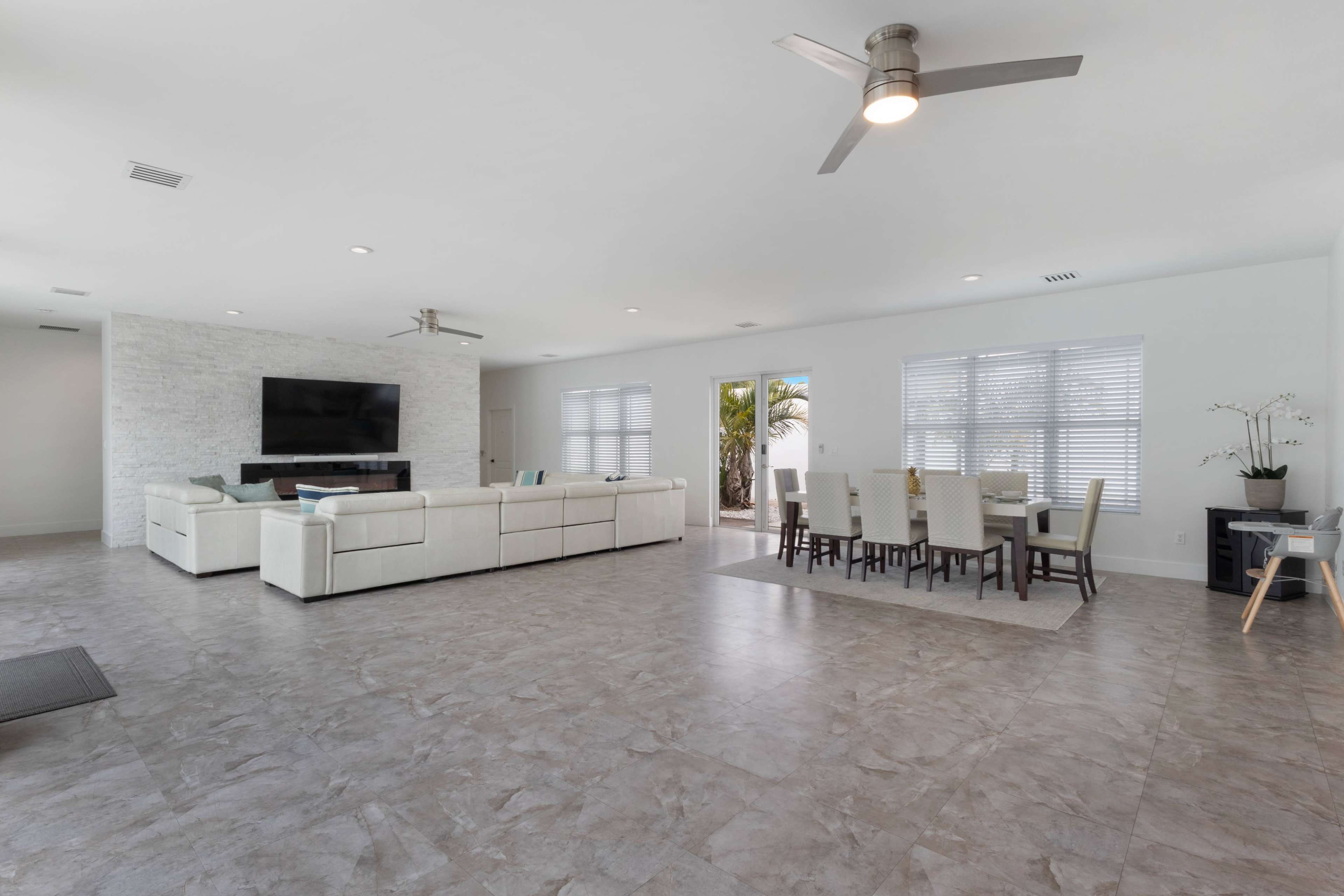 The image depicts a spacious living area with a sectional sofa, a wall-mounted television, and a dining table set for six near large windows.