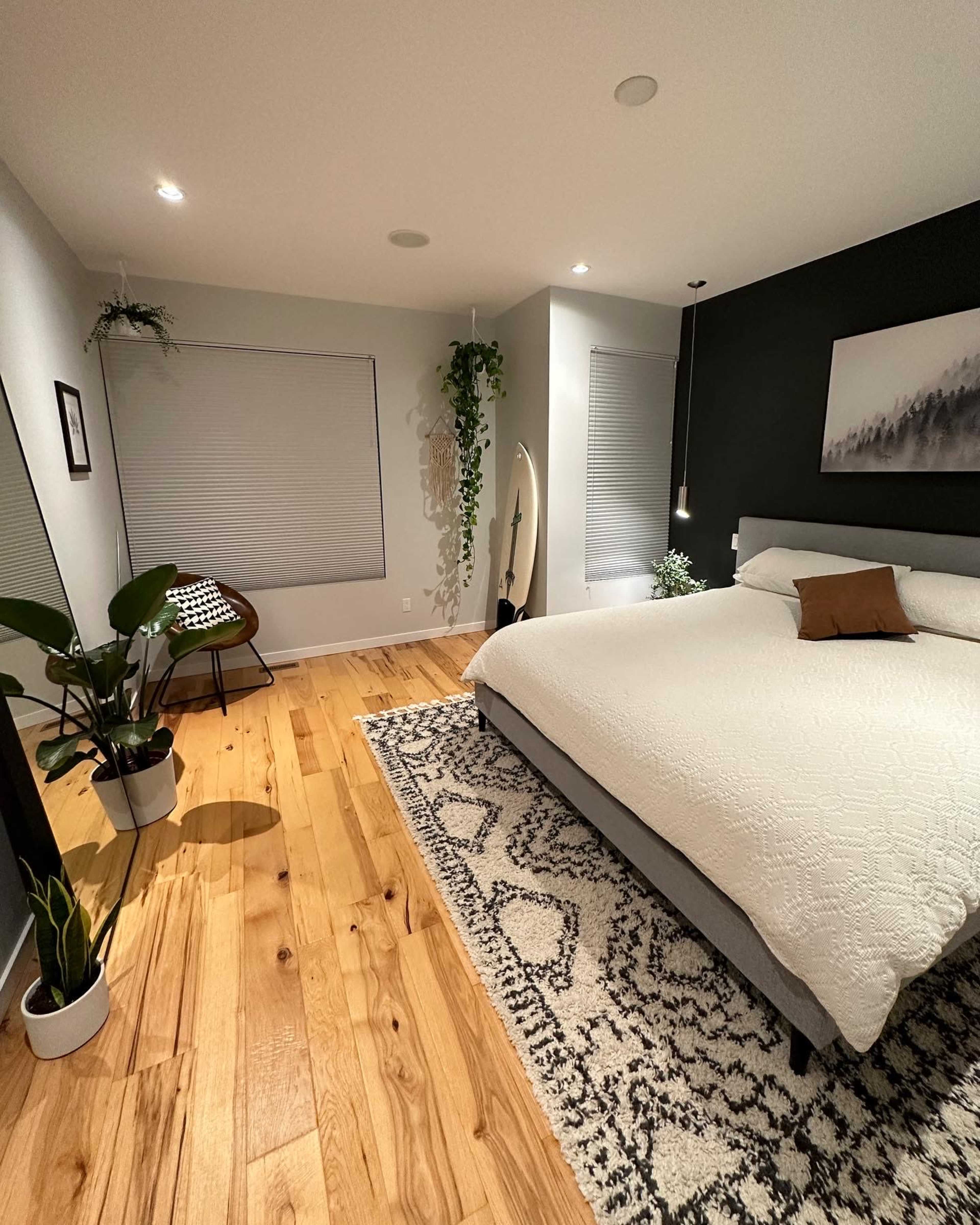 The bedroom features a large bed with a white coverlet, a patterned rug on wooden flooring, a chair in the corner, and plants against a dark wall.
