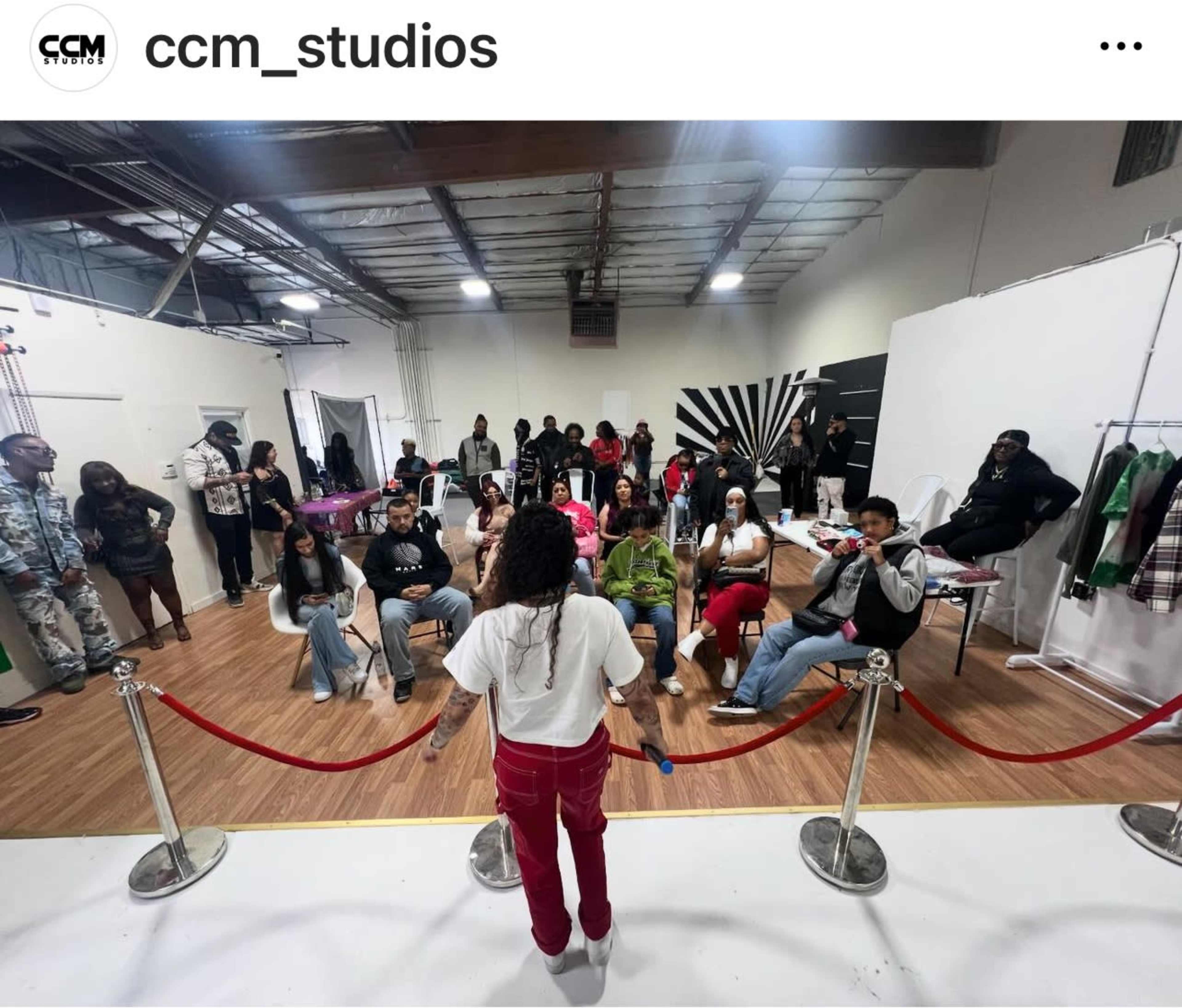 A speaker stands in front of a seated audience in a studio setting, with a red rope barrier separating them.