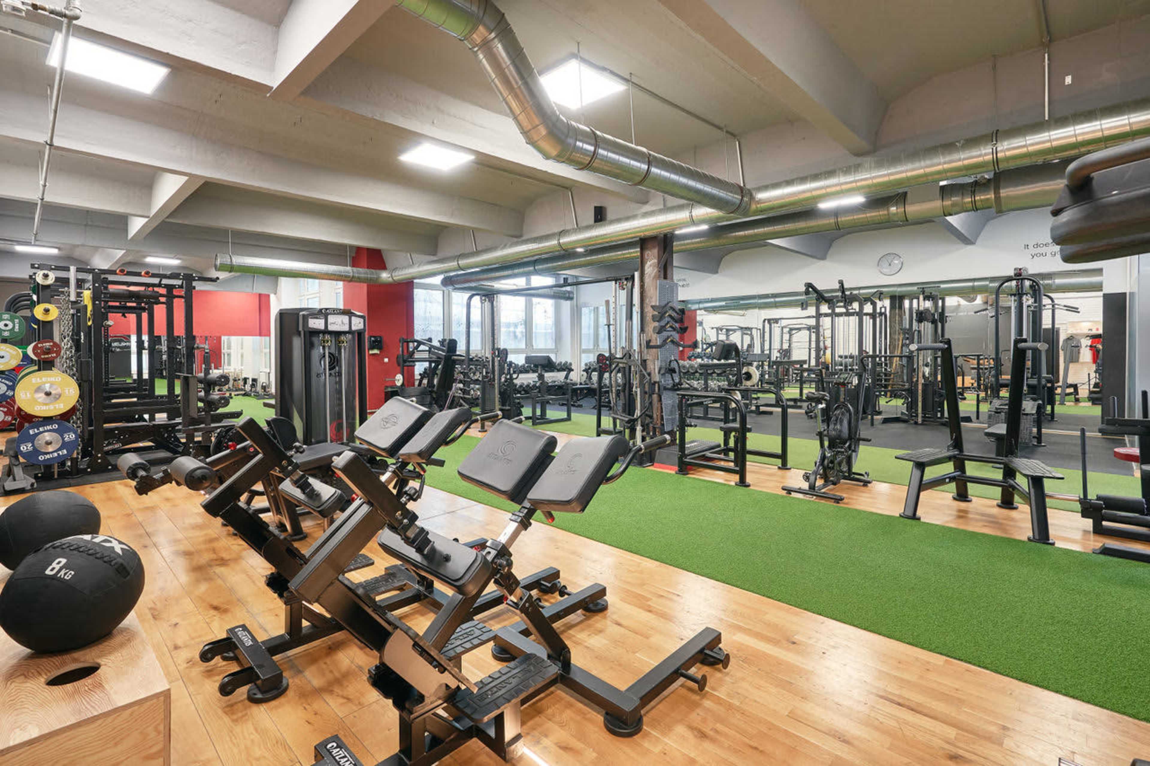 The image shows a spacious gym interior featuring various weightlifting equipment arranged on green carpet and wooden flooring.