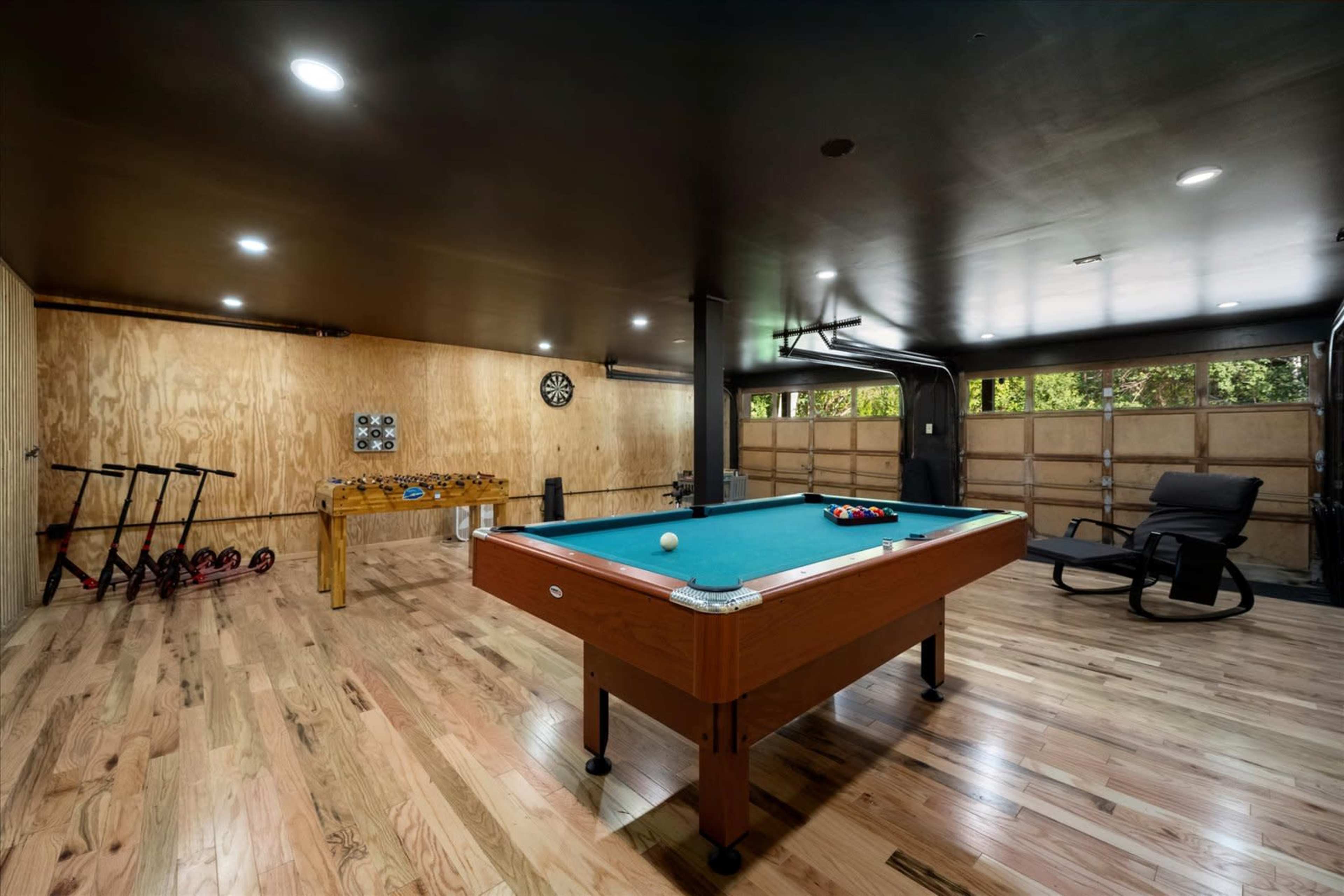 The image shows a game room with a pool table, a dartboard on the wall, and large garage doors that open to a garden area.