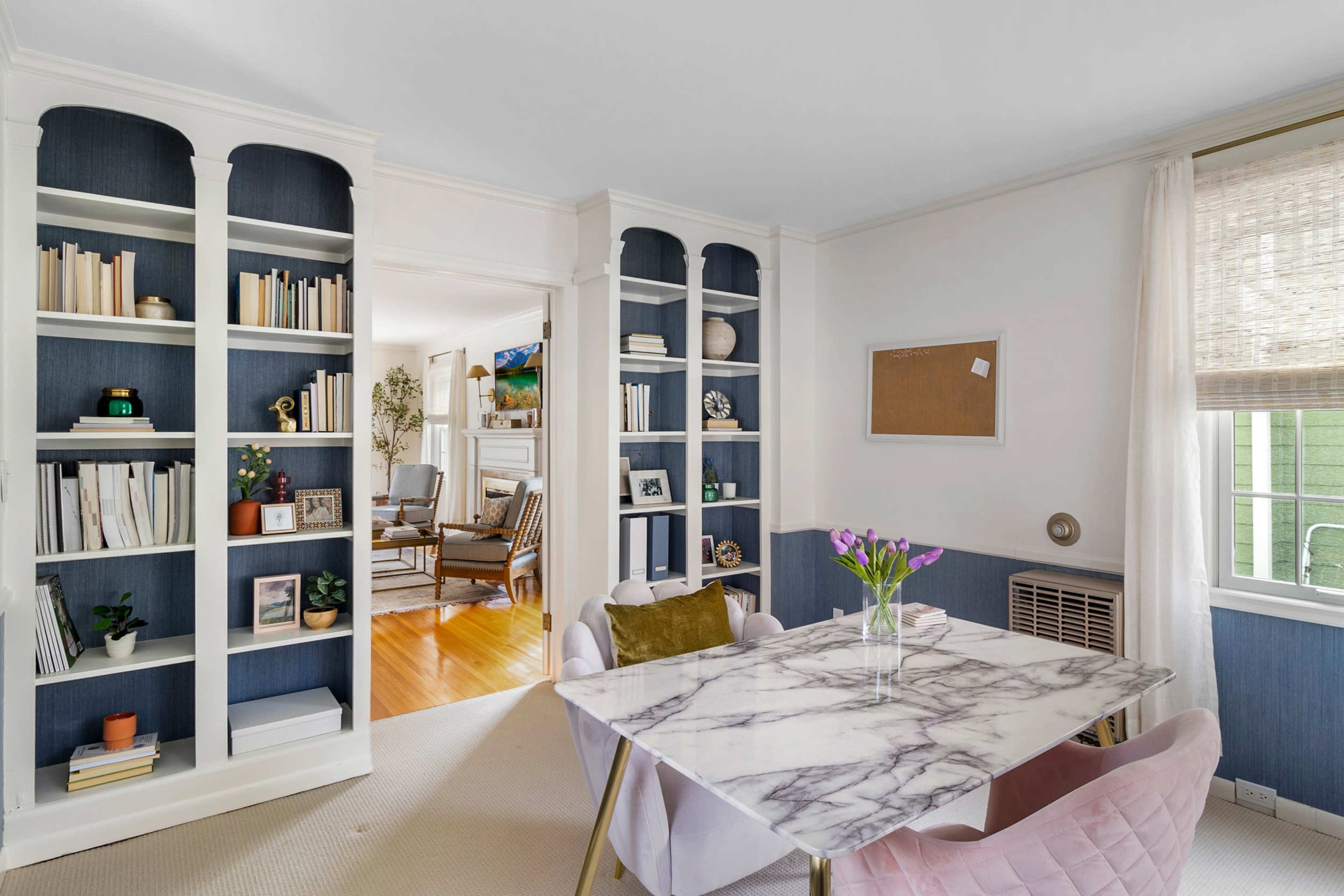 A dining area features a marble-topped table surrounded by pink chairs, with built-in bookshelves displaying books and decorative items on one side.