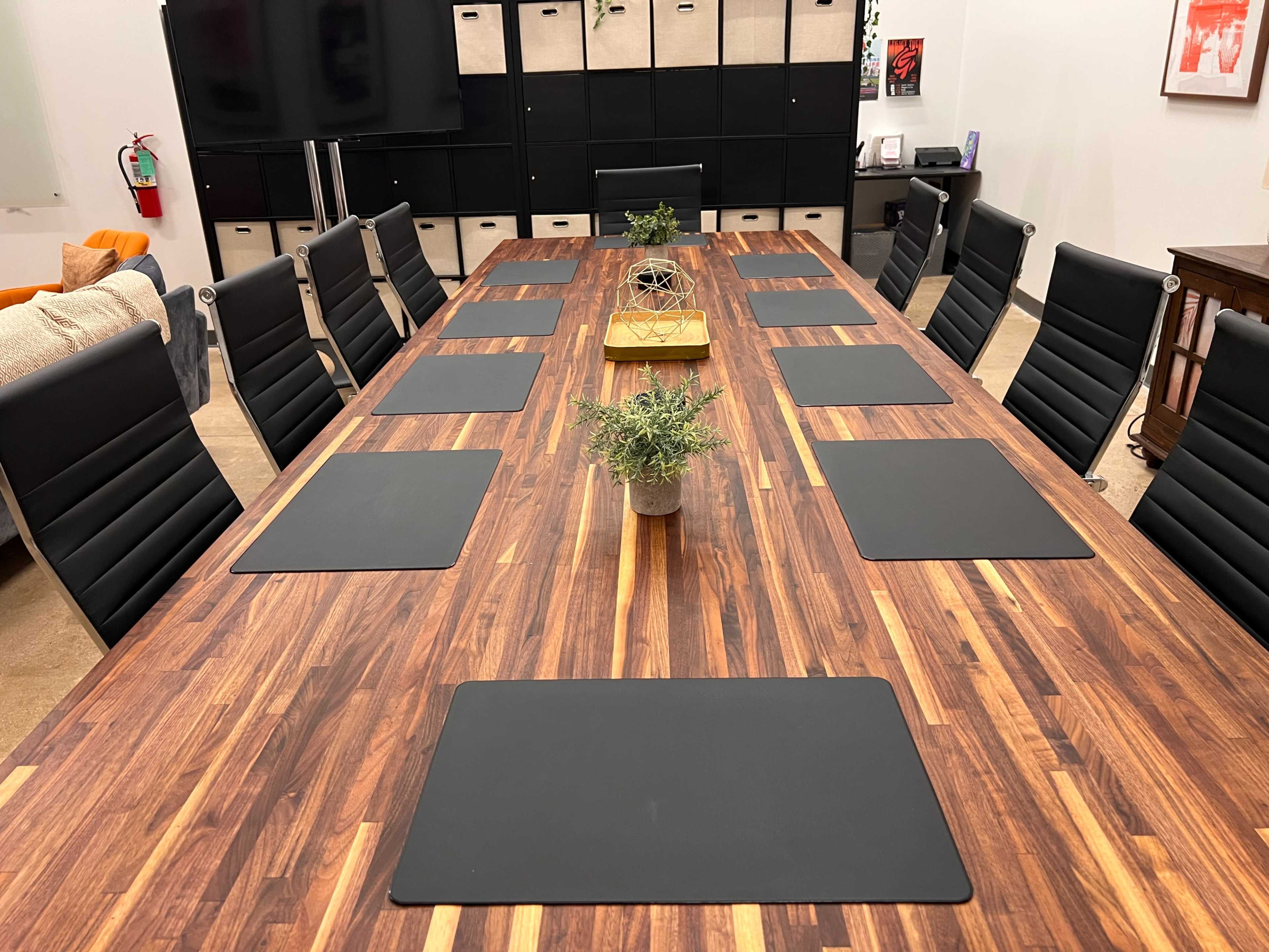 A long wooden conference table is lined with black chairs, placemats, and small potted plants, in a modern meeting room with storage cabinets in the background.