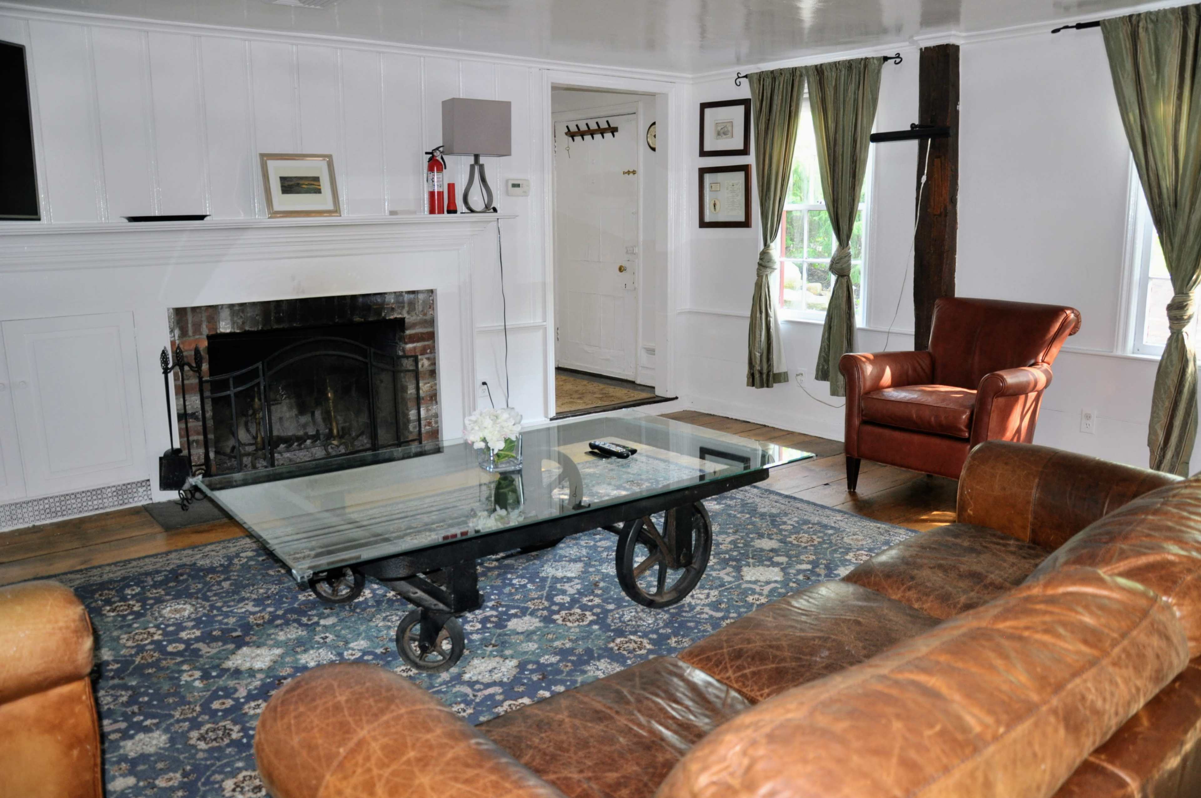 The living room features a glass coffee table on wheels, a brown leather sofa, an armchair, and a fireplace flanked by windows with curtains.
