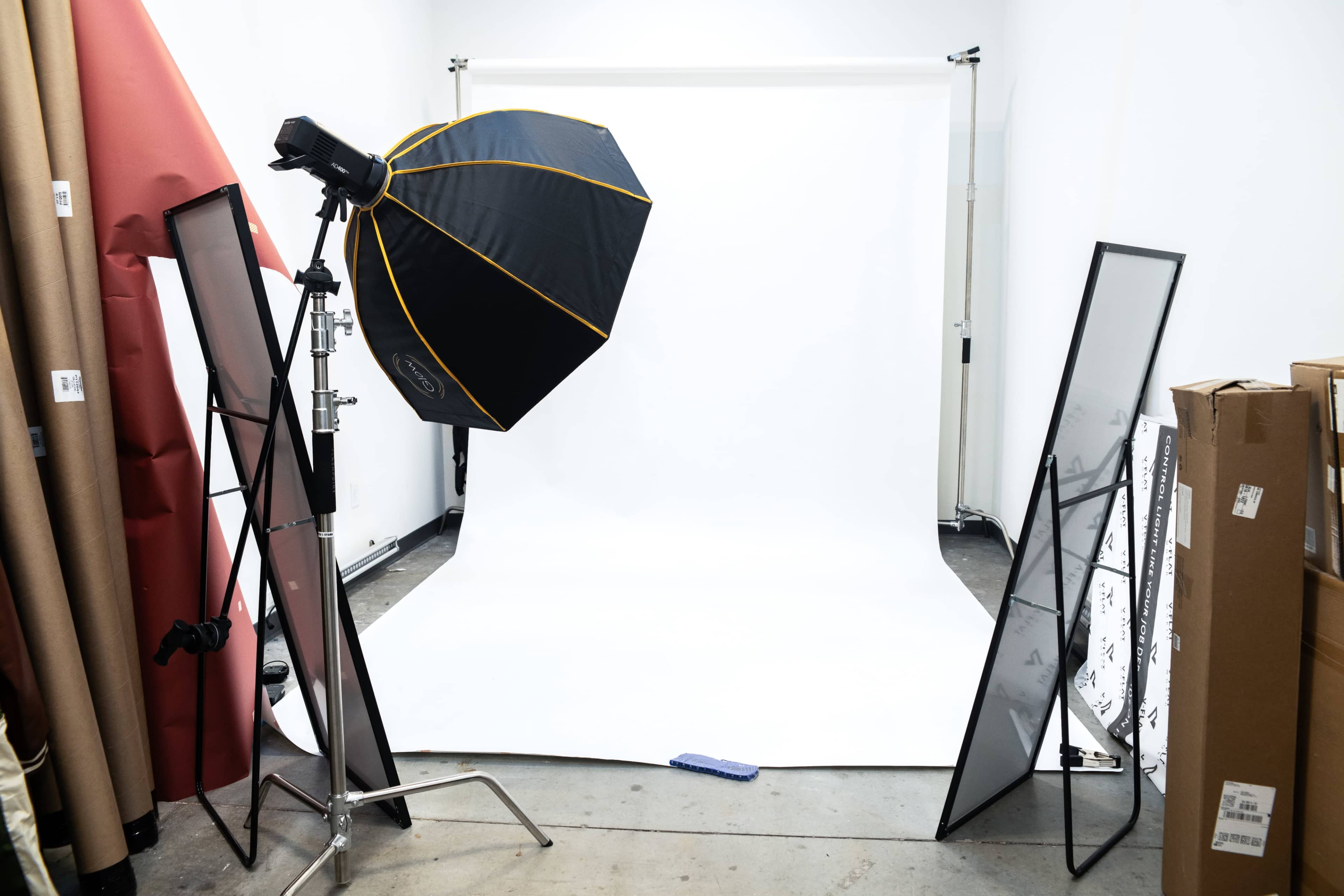 The image shows a photography studio setup with a large softbox light positioned beside a white backdrop and various props, including mirrors and rolls of paper, stored against the walls.