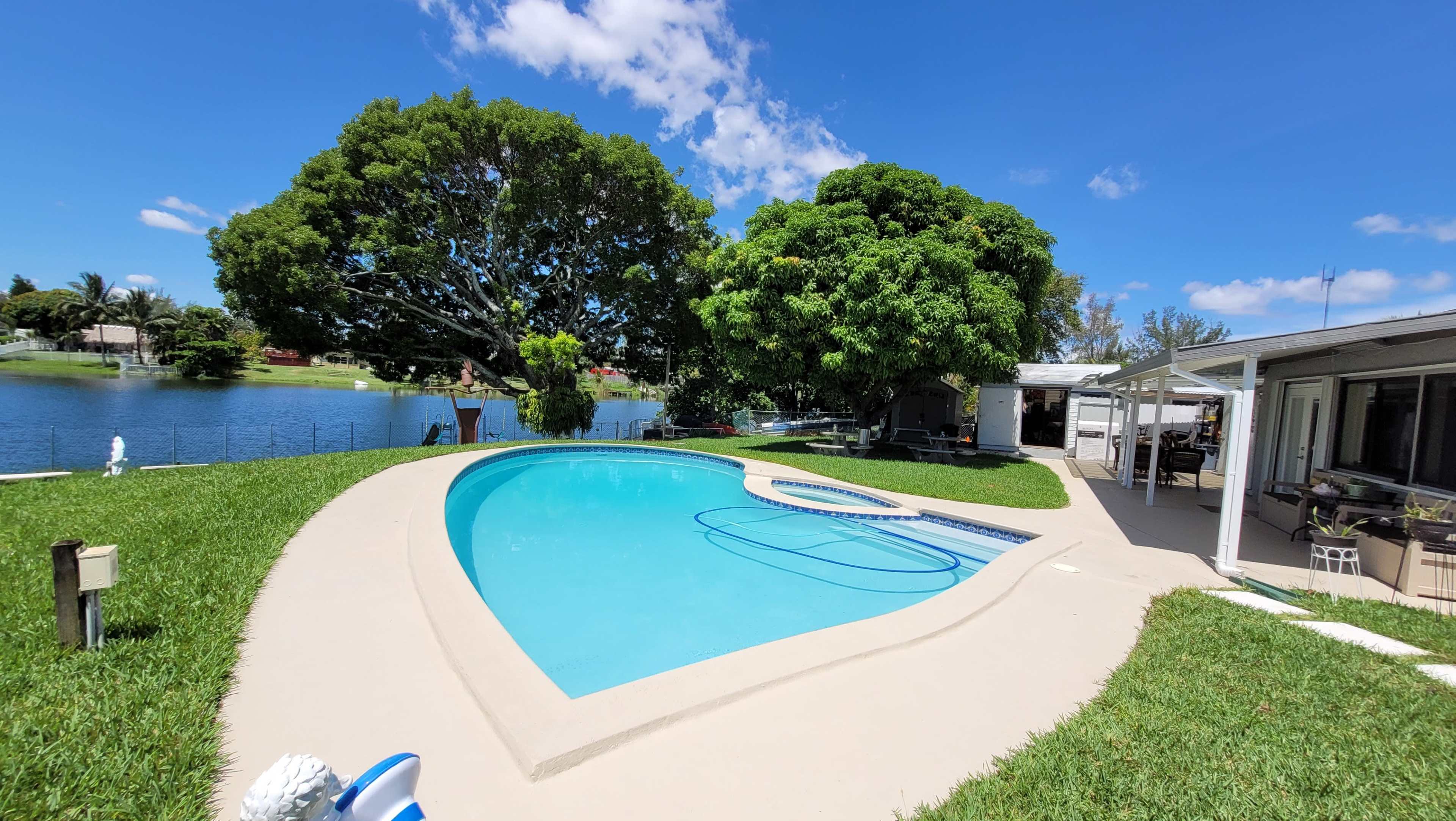 A swimming pool surrounded by grass and large trees, with a view of a lake and a house in the background.