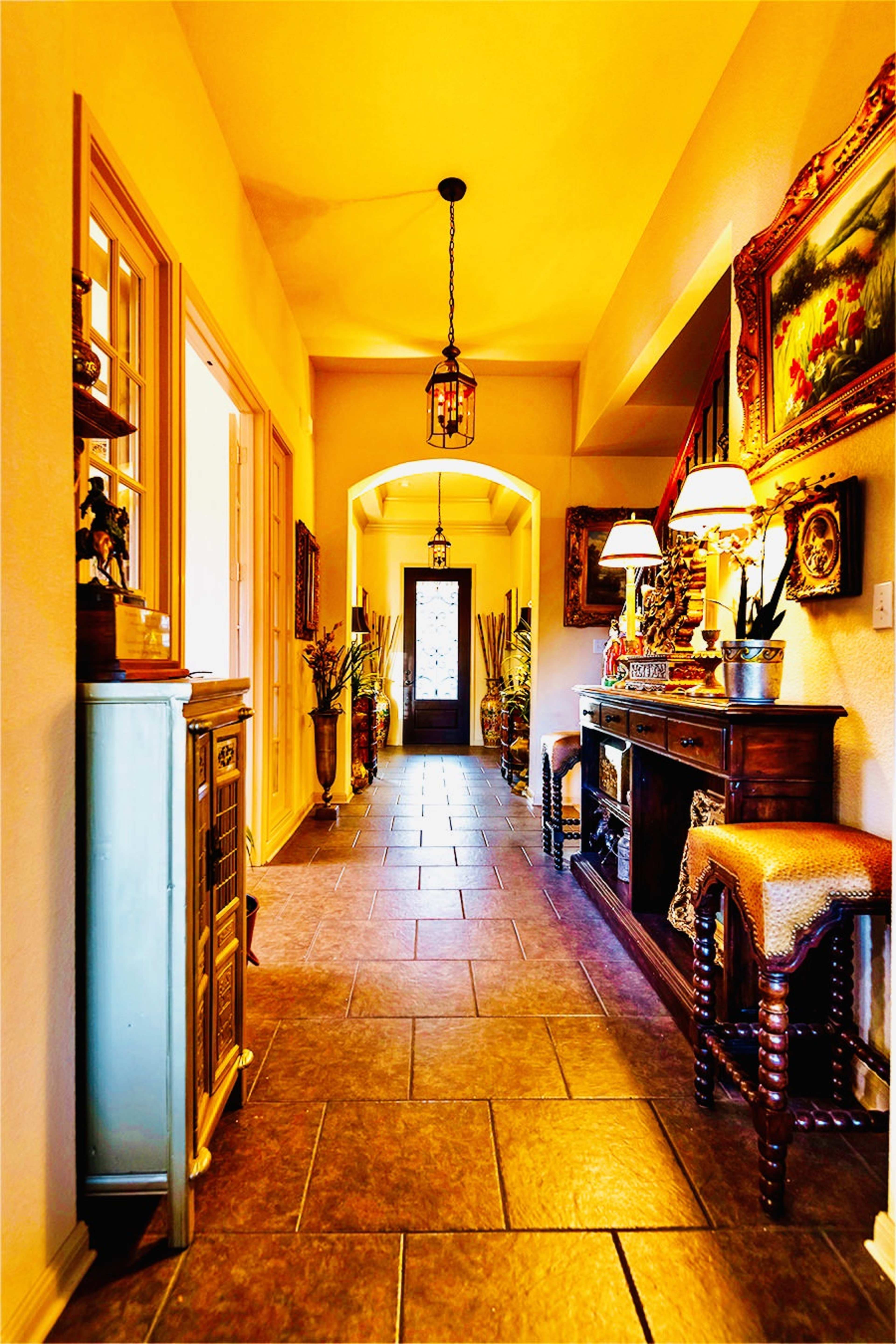 A brightly lit hallway with a tiled floor, decorated with various plants, artwork, and furniture on each side.