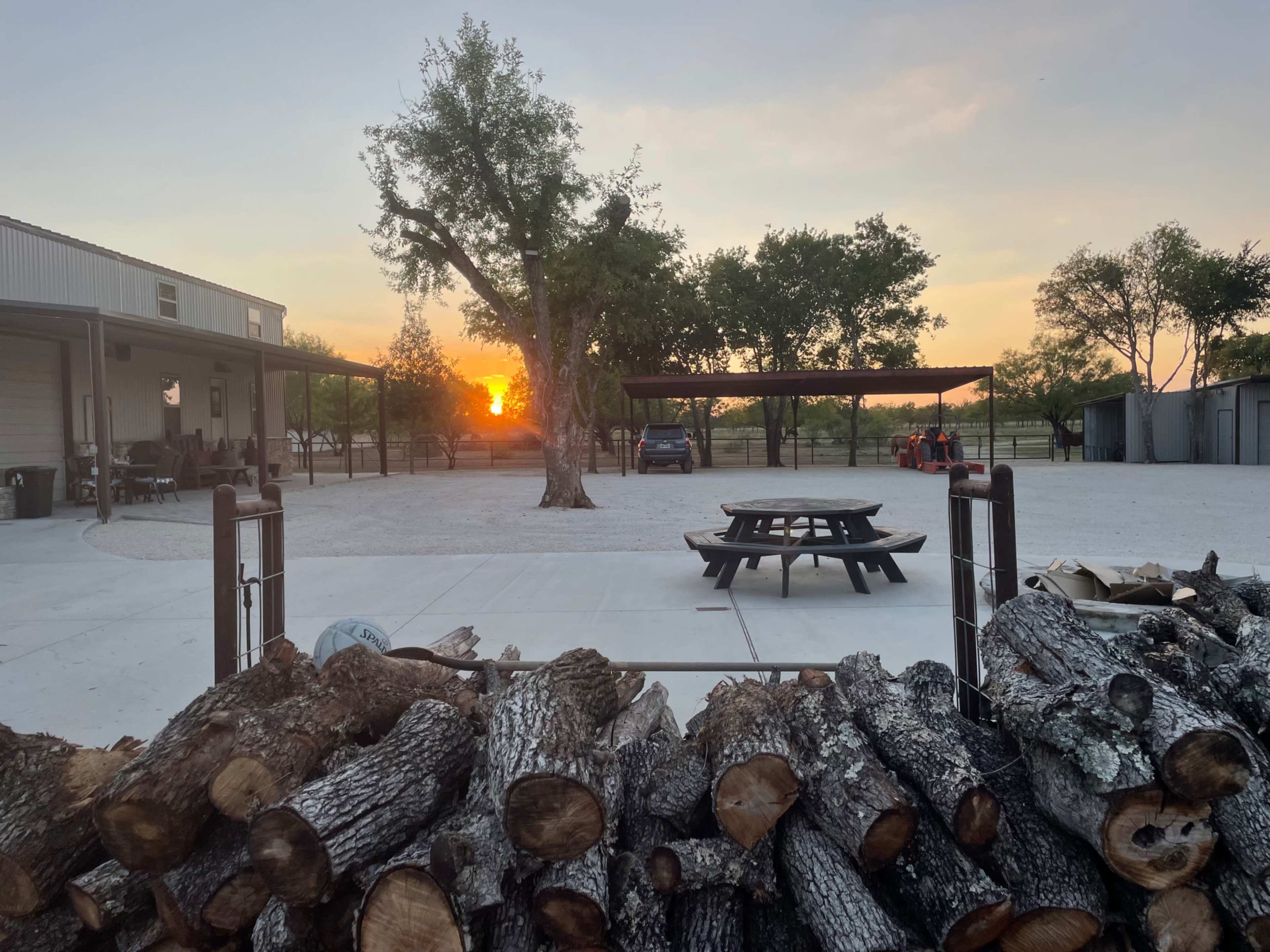 The image shows a stack of firewood in the foreground with a gravel area and a picnic table, while the sun sets behind trees and a building in the background.