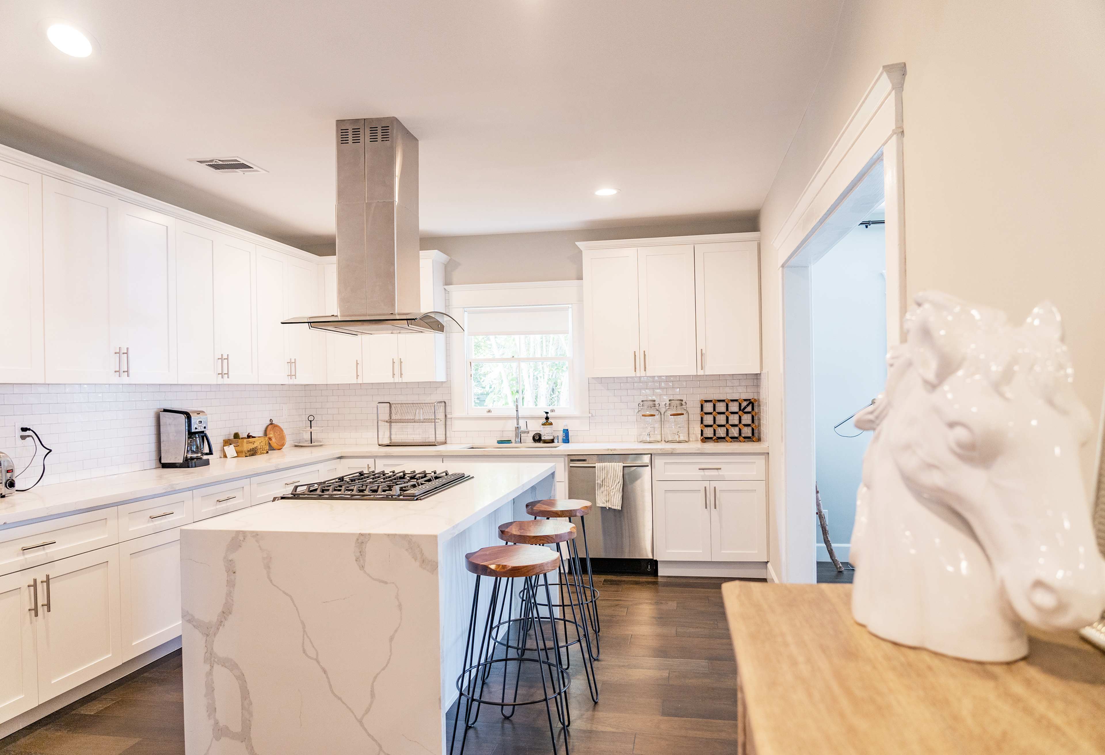 A modern kitchen features white cabinets, a central island with a gas cooktop, and barstools, illuminated by natural light from a nearby window.