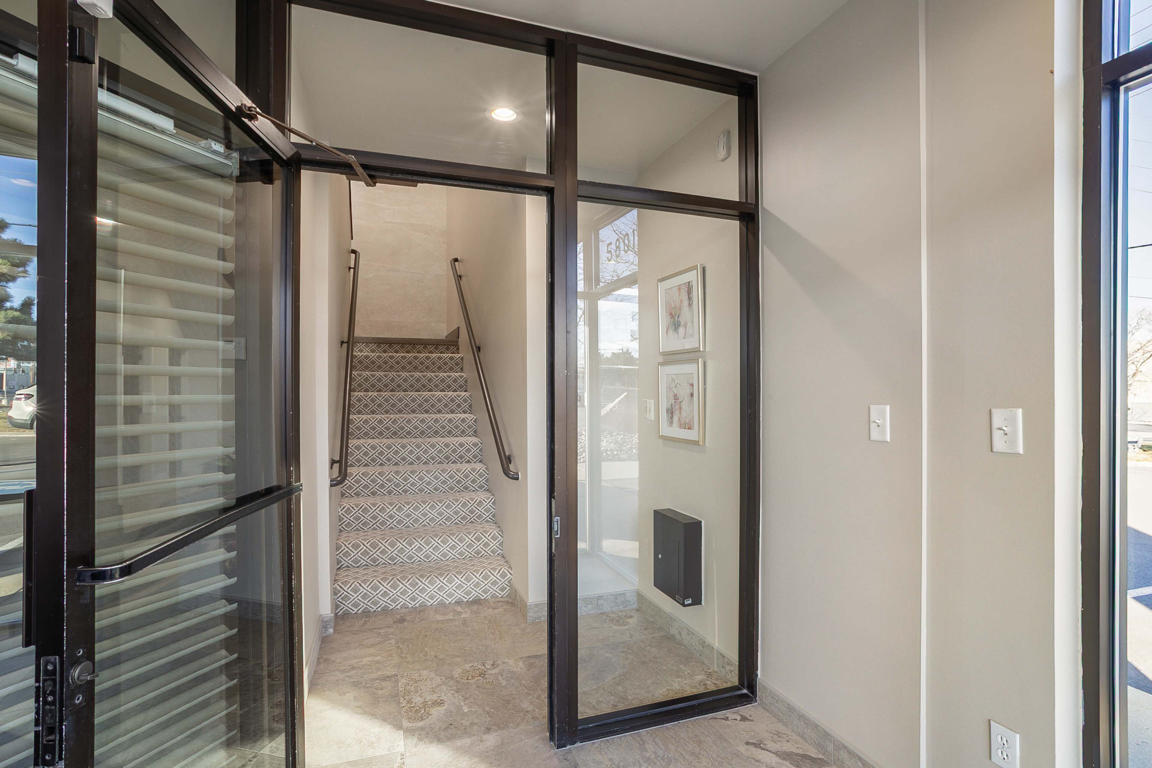 The image shows an entrance area featuring a glass door leading to a staircase with patterned steps and framed artwork on the wall.