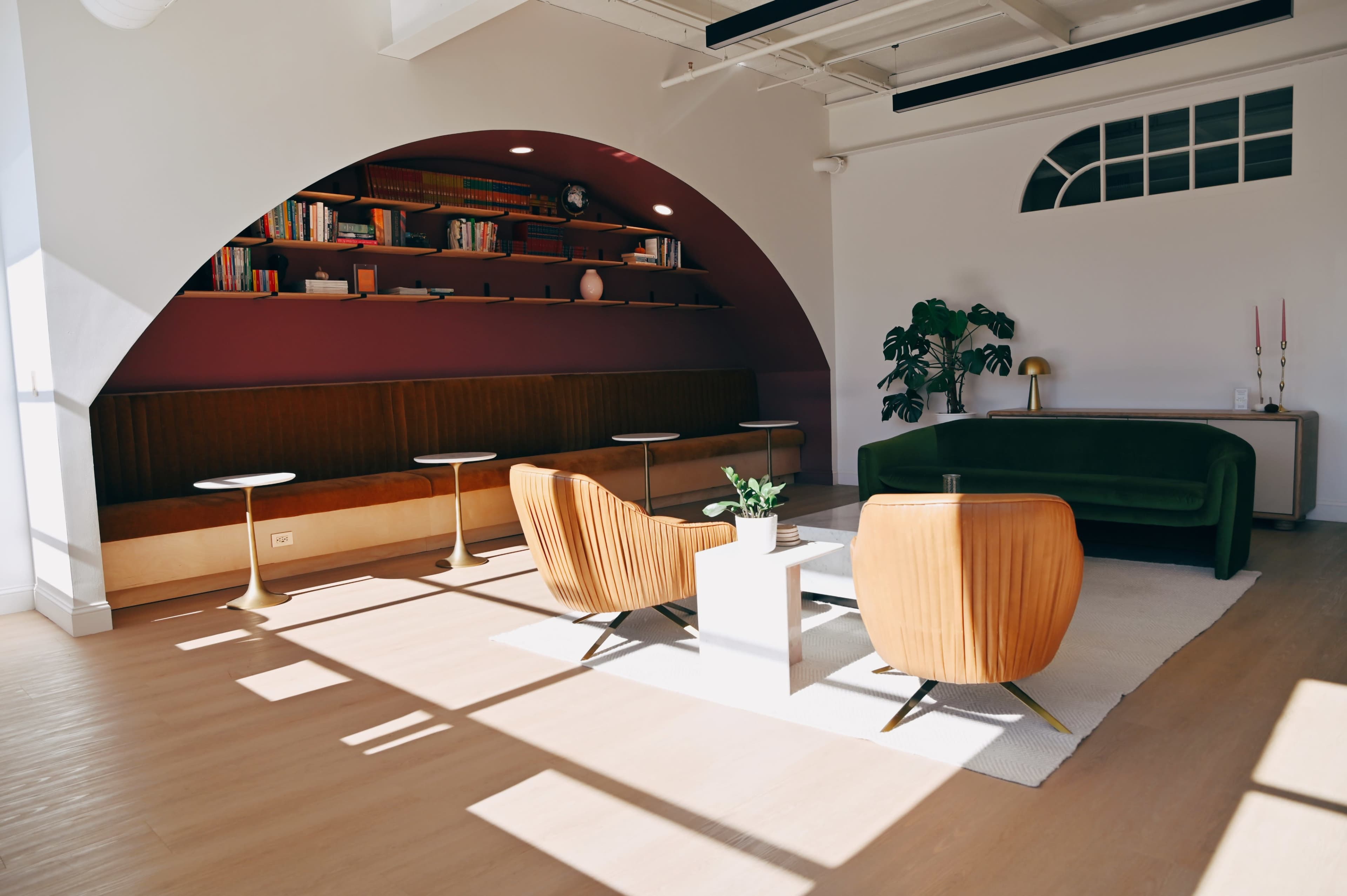 The image shows a modern lounge area with curved architecture, featuring a seating space with bookshelves, two armchairs, a small table, and large windows allowing natural light to flood the room.