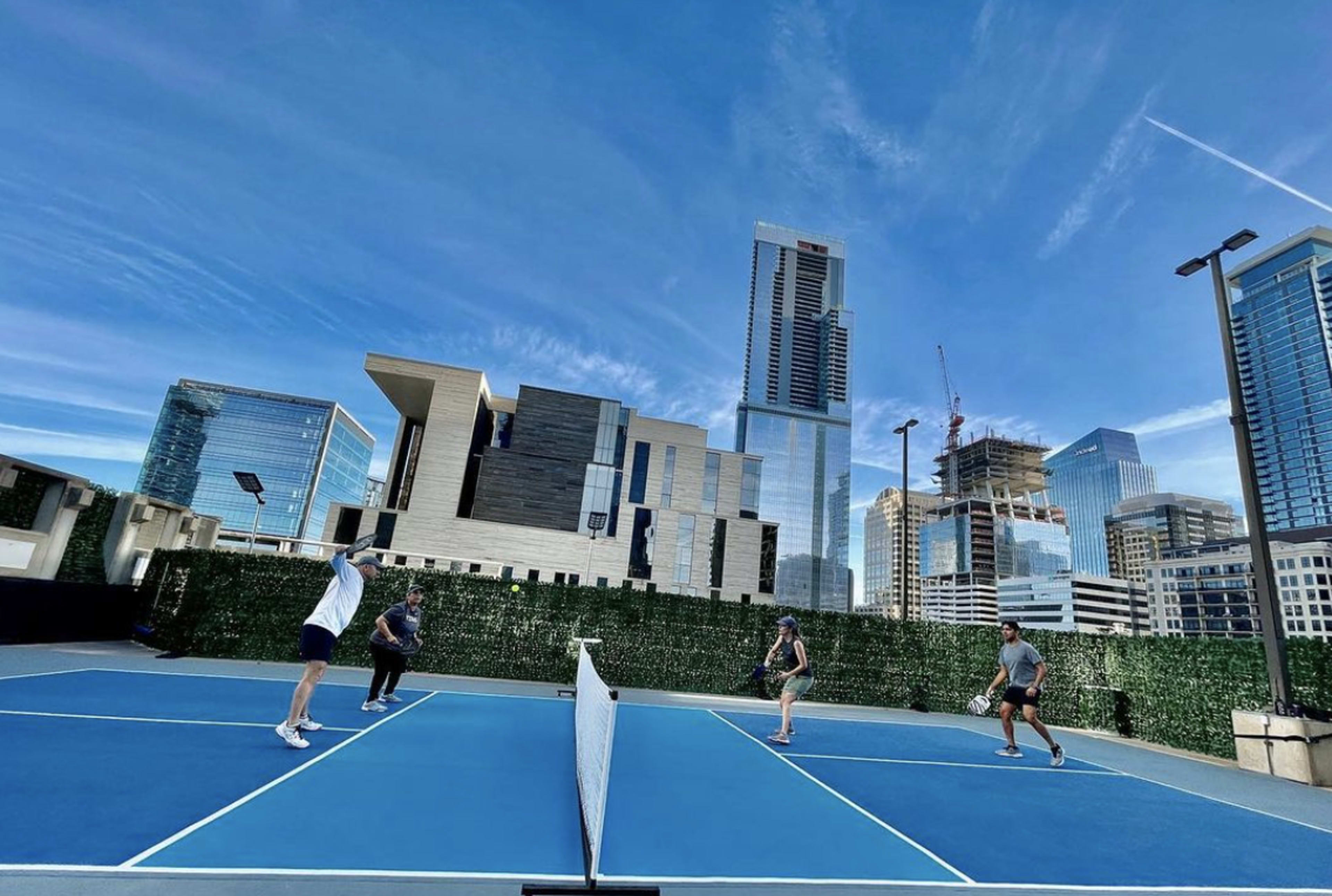 Four people play tennis on a blue court surrounded by tall buildings under a clear sky.