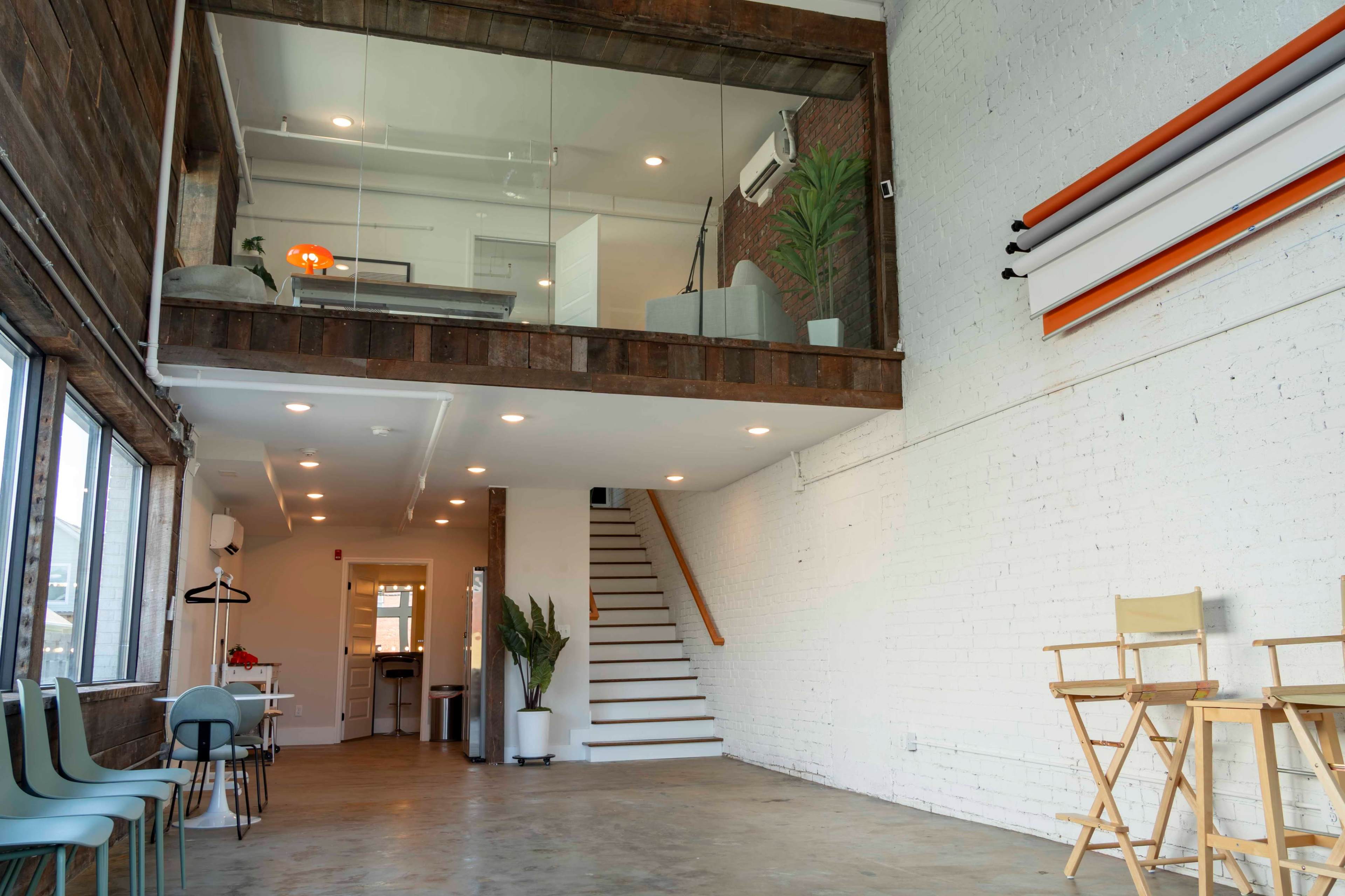 The image shows a spacious interior with a multi-level design featuring wooden accents, a staircase, and seating areas, illuminated by natural light from large windows.