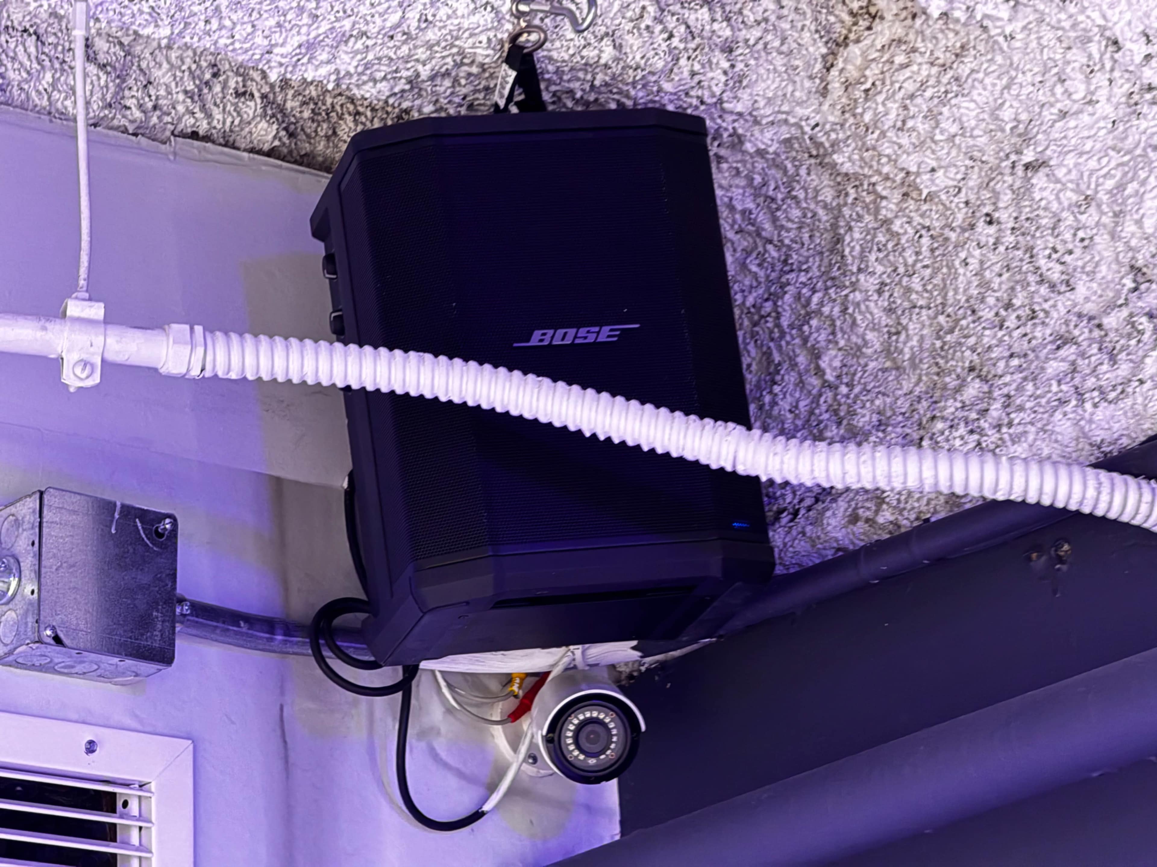 A black Bose speaker is mounted on the ceiling, with white piping and a security camera nearby.