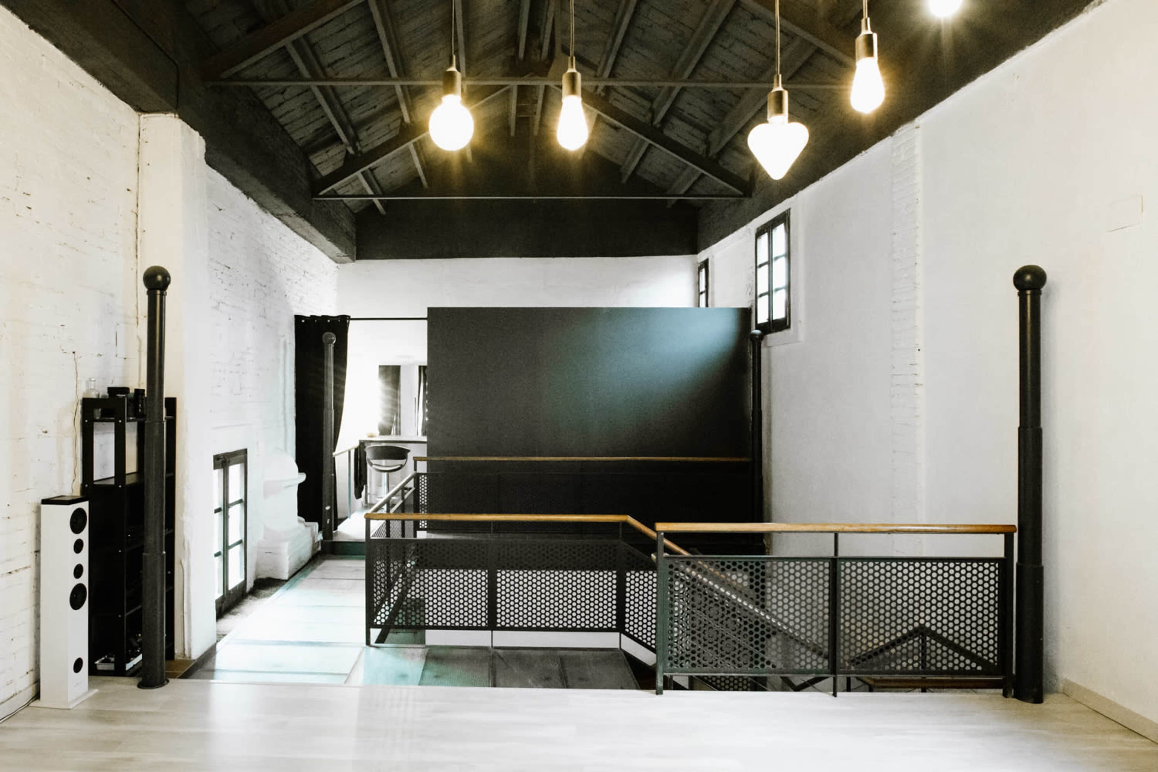 A modern interior space with high ceilings, wooden flooring, and an upper level overlooking a lower area, featuring large windows and industrial-style lighting.