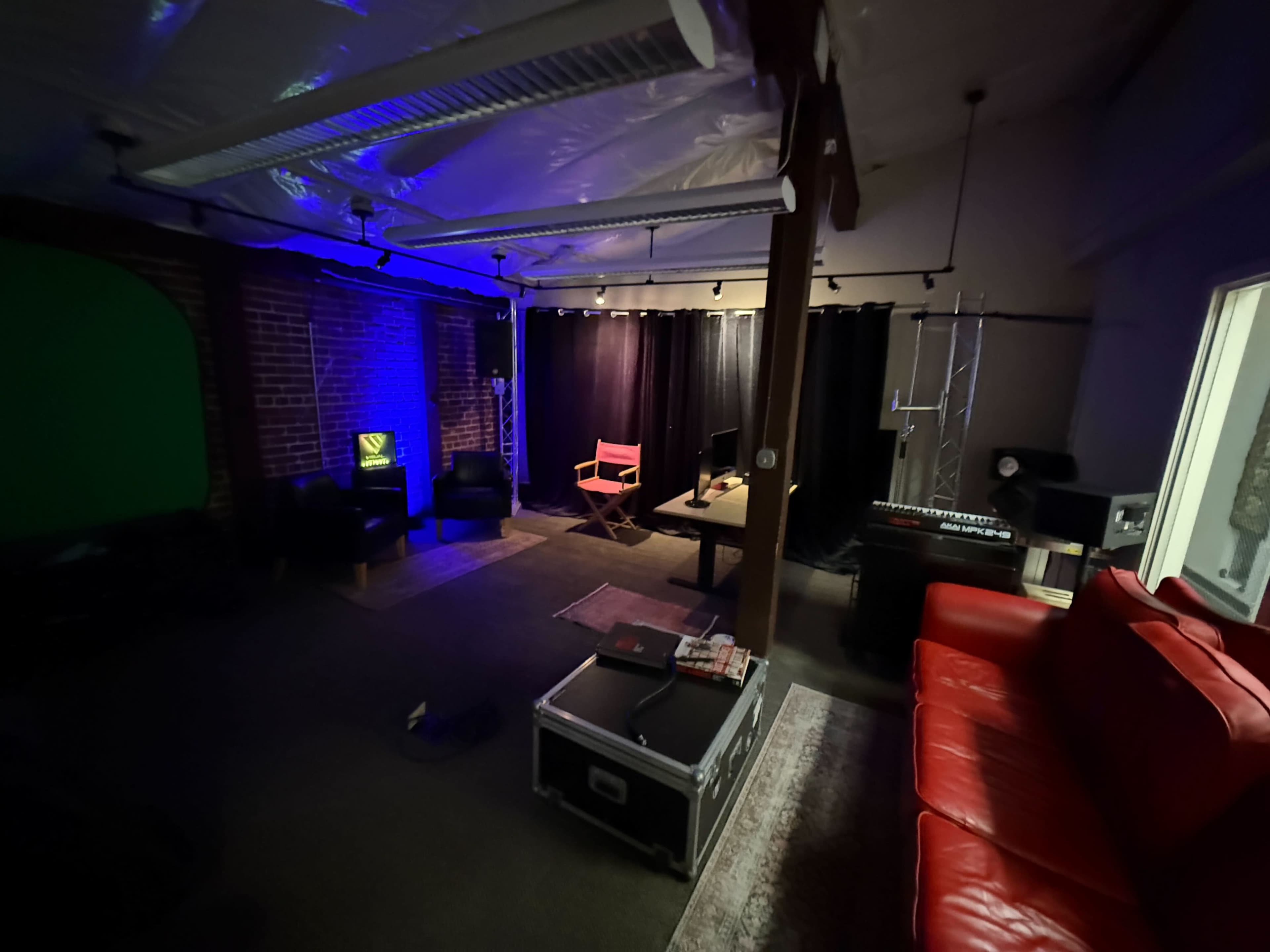 The image shows a dimly lit room with a red couch, a workspace with a desk and computer, and various seating areas, complemented by purple and green ambient lighting.