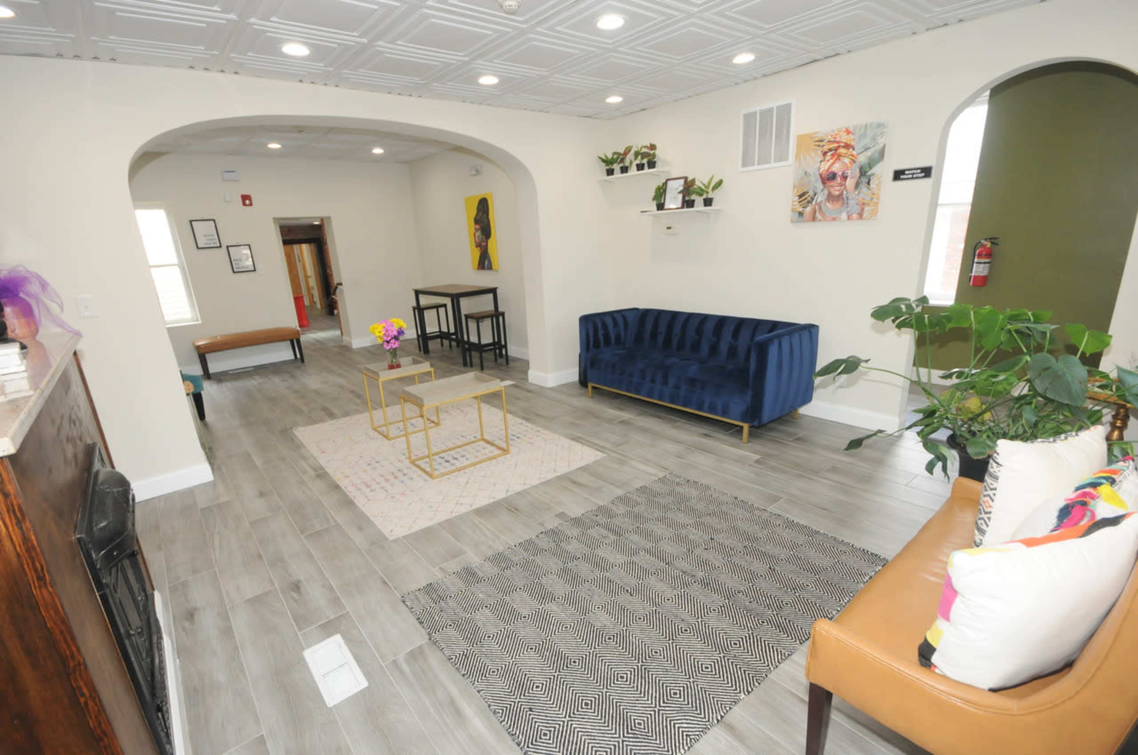 The image shows a spacious lobby area with a blue sofa, a brown couch, coffee tables, and decorative rugs, illuminated by natural light.