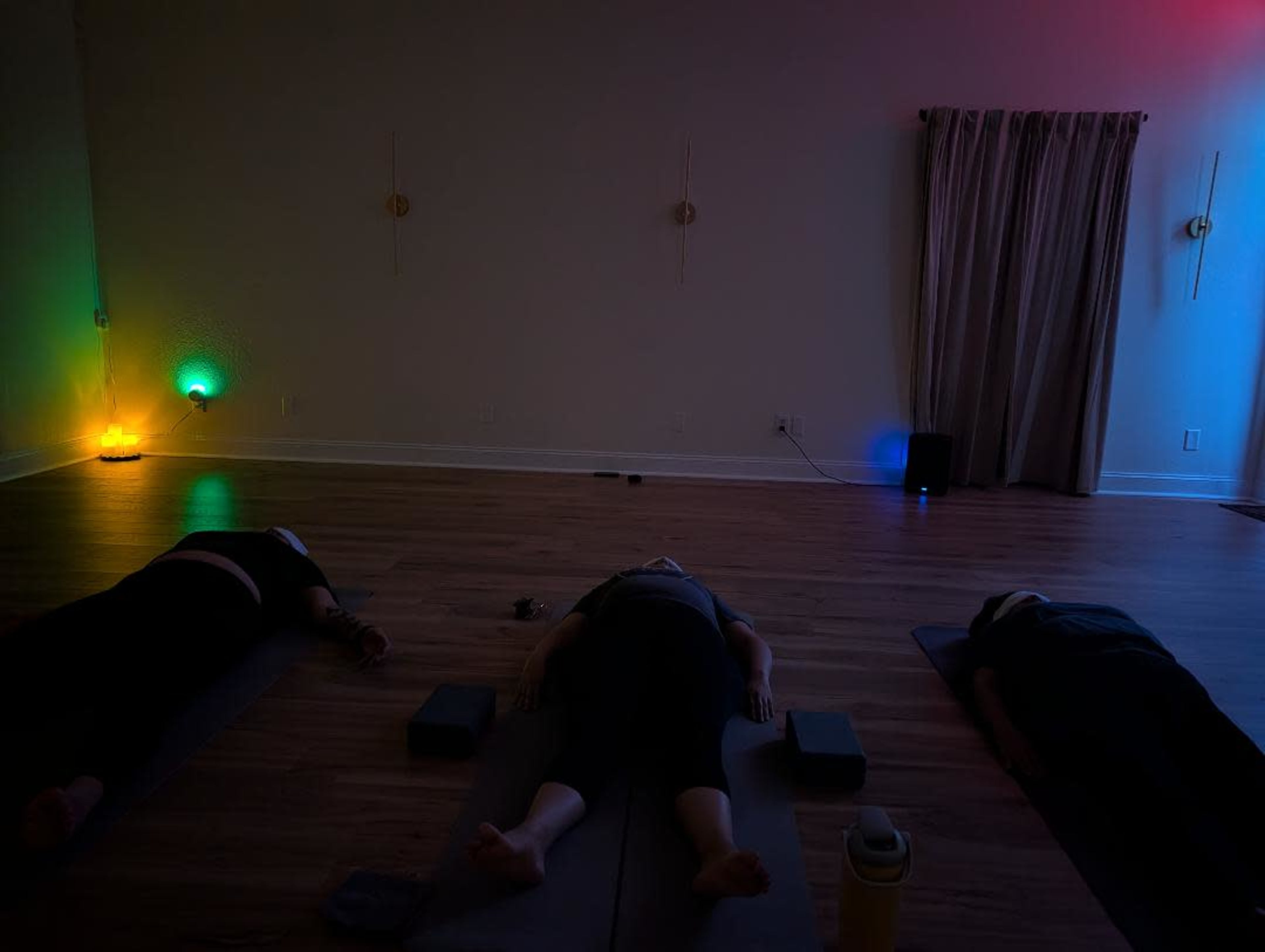 Three individuals are lying on yoga mats in a softly lit room, with colored lights illuminating the space.