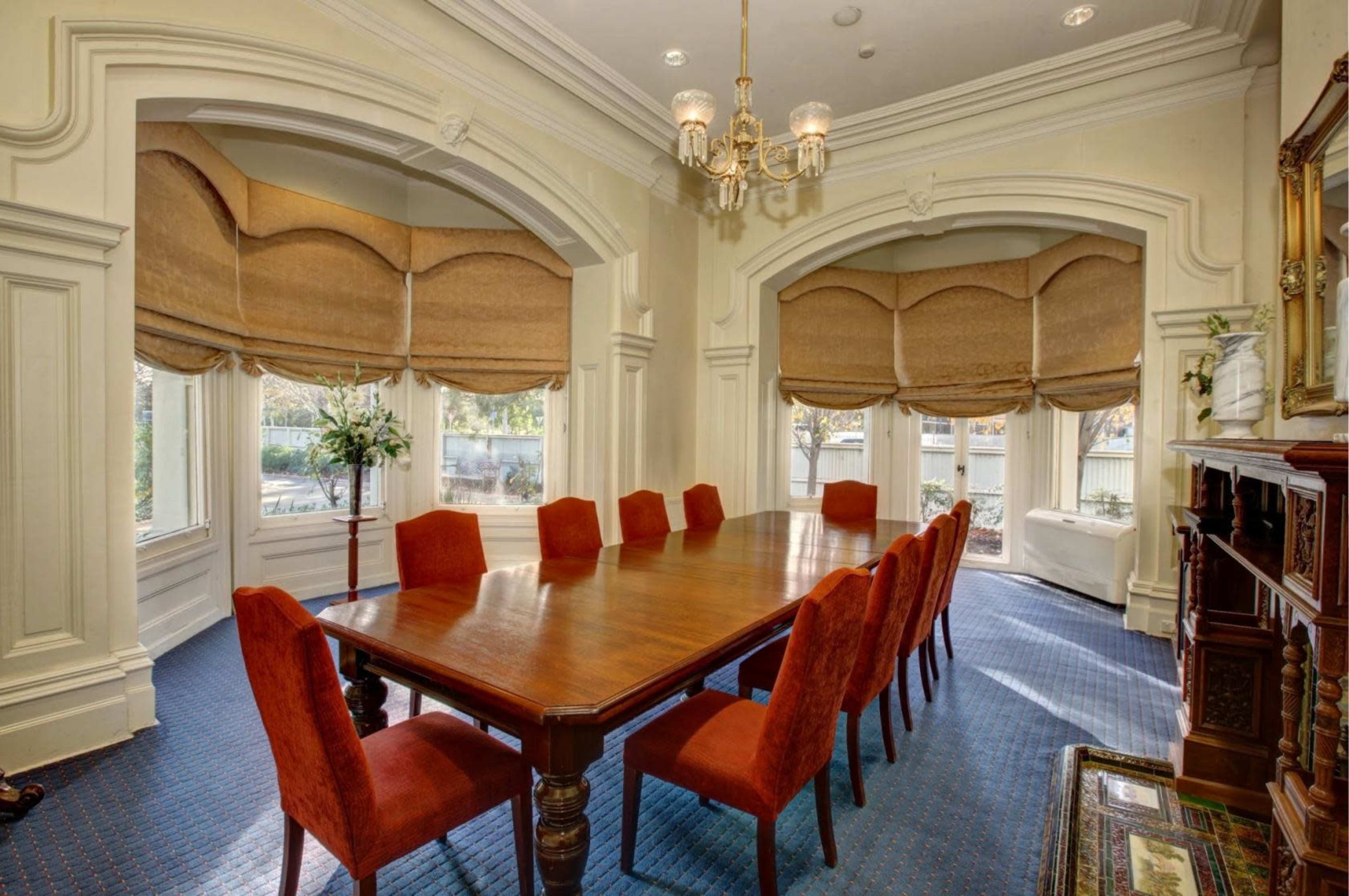 A long wooden dining table surrounded by red upholstered chairs is positioned in a well-lit room with large windows and decorative window treatments.