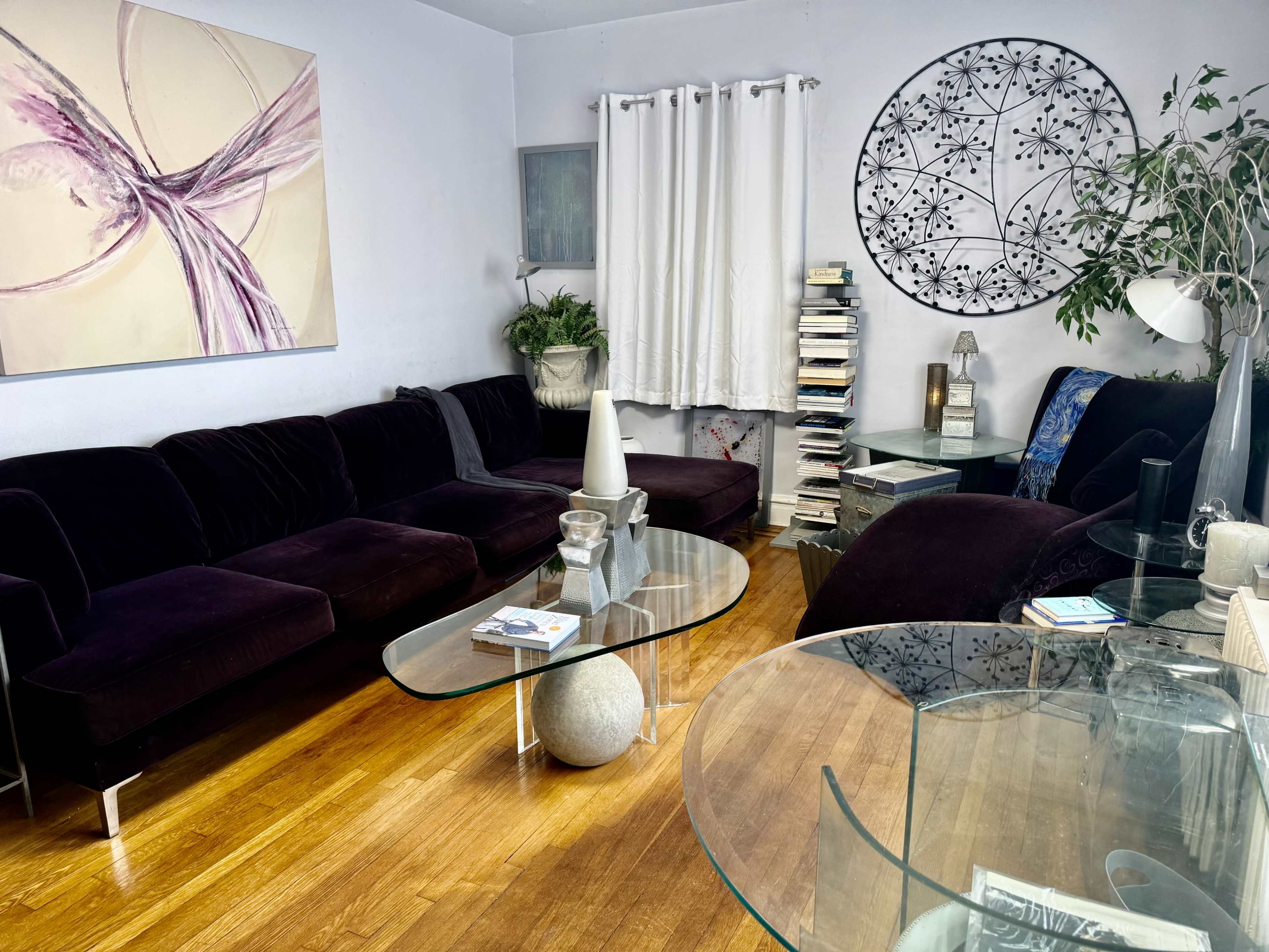 The image shows a cozy living room with a dark velvet sectional sofa, a round wall art piece, and glass tables atop wooden flooring.
