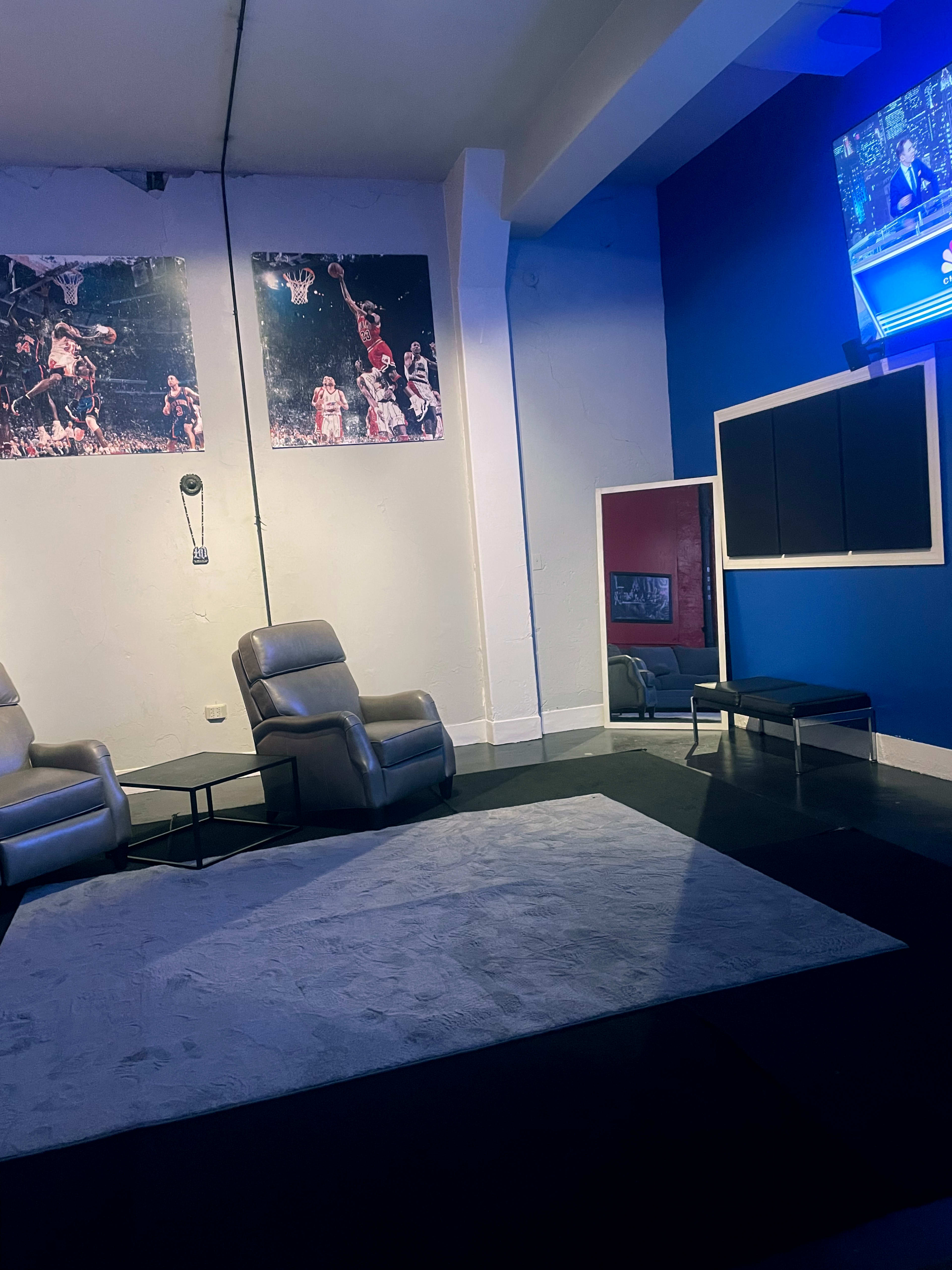 The image shows a lounge area with two reclining chairs, a low table, a rug, and wall-mounted basketball-themed artwork, along with a television displaying a sports broadcast.
