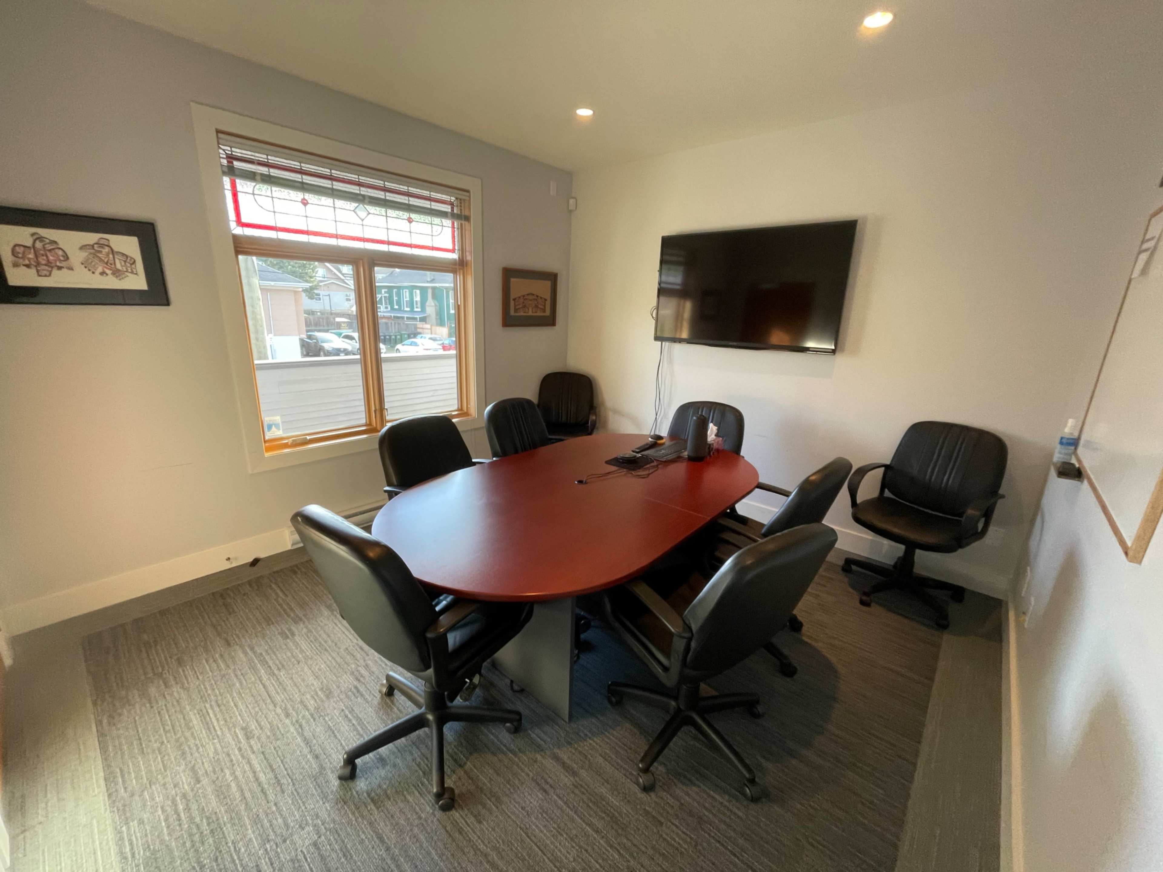 A conference room with a large oval table surrounded by six black chairs and a wall-mounted television.