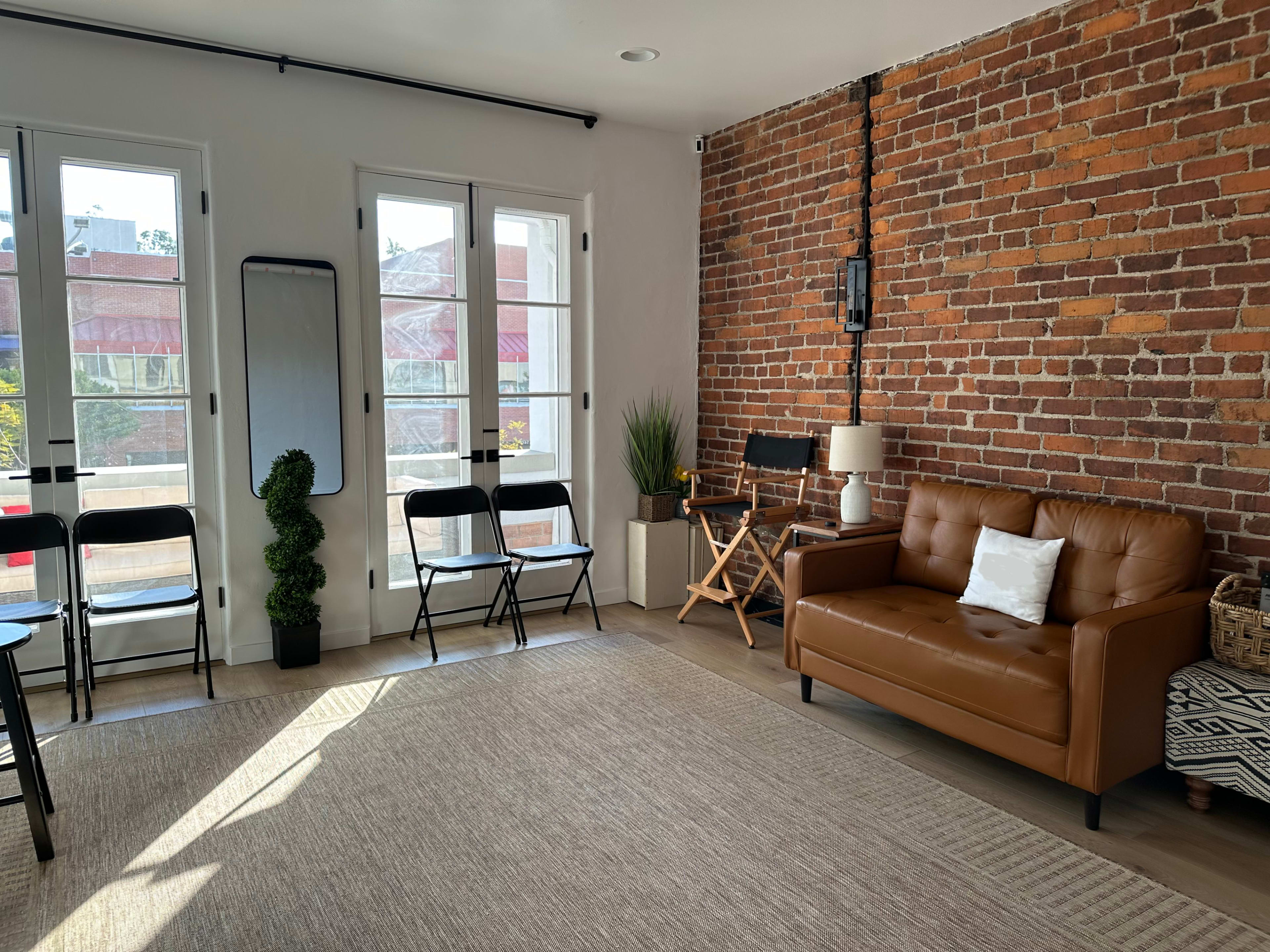 The image shows a room with exposed brick walls, featuring a leather couch, a chair, a potted plant, and several black folding chairs arranged along the wall beside large windows.