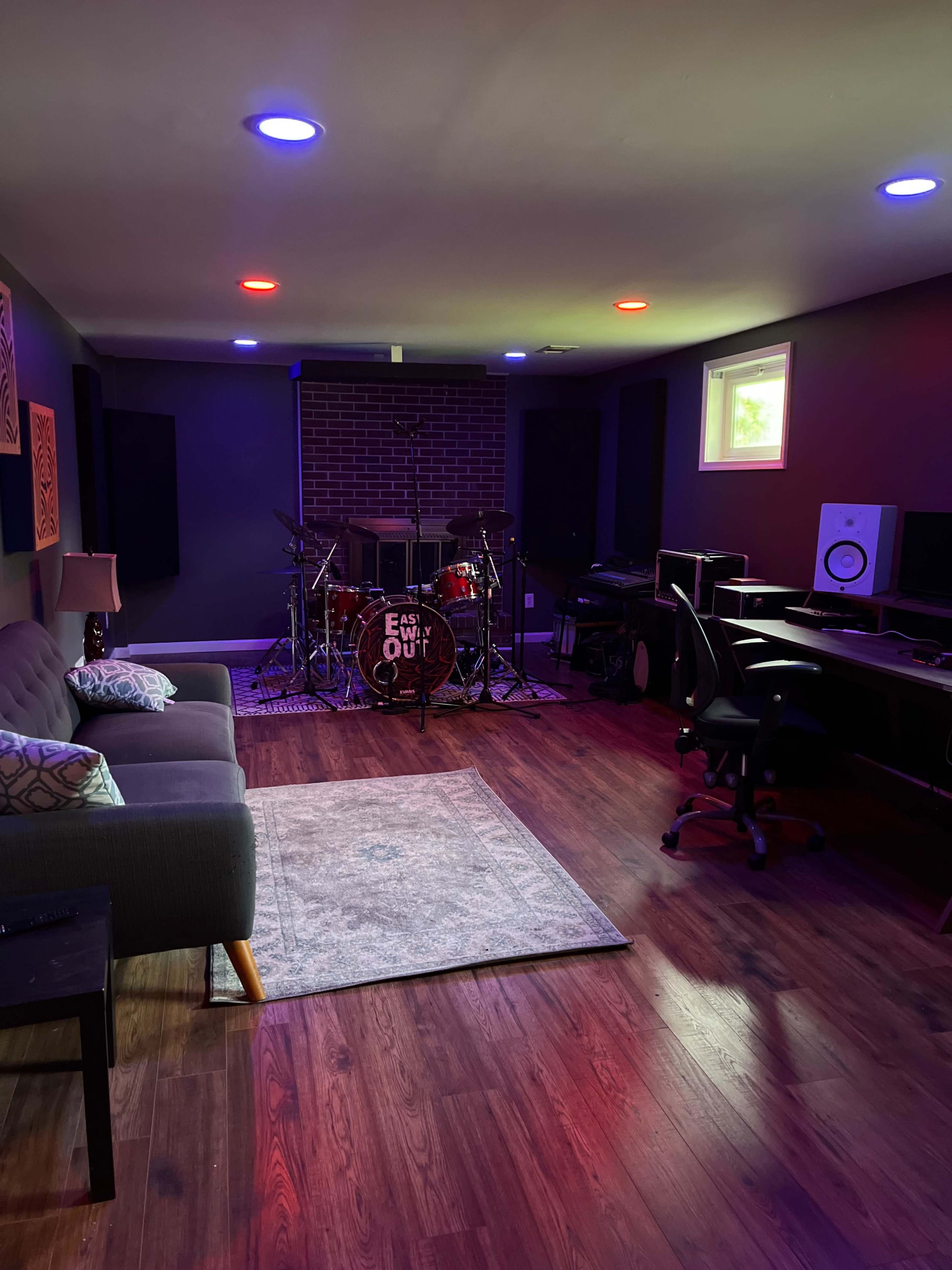 A music studio featuring a drum set in front of a brick wall, with a seating area and workspace equipped with monitors and a desk.