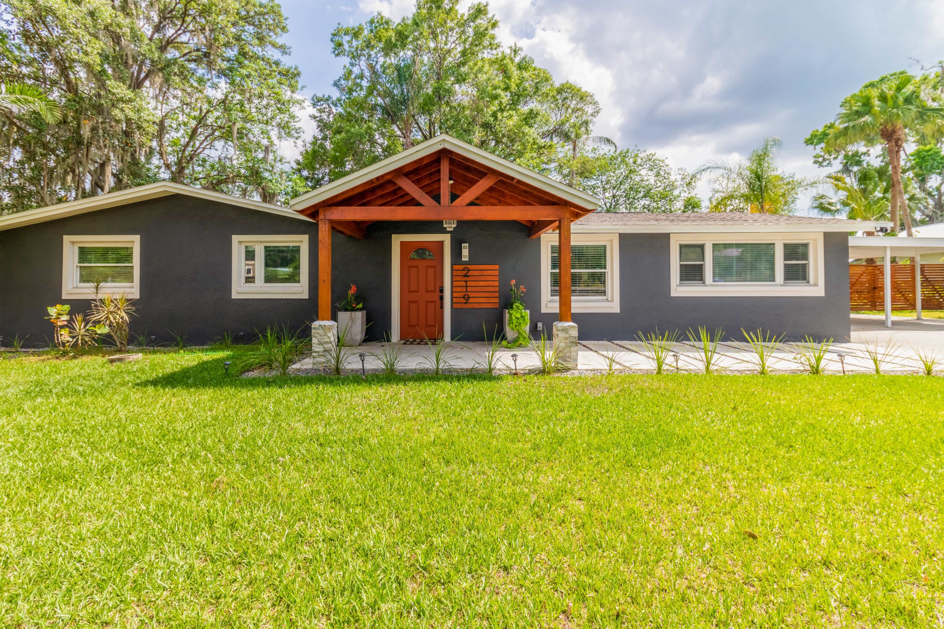 A single-story house with a wooden entry awning and landscaped front yard is surrounded by trees.