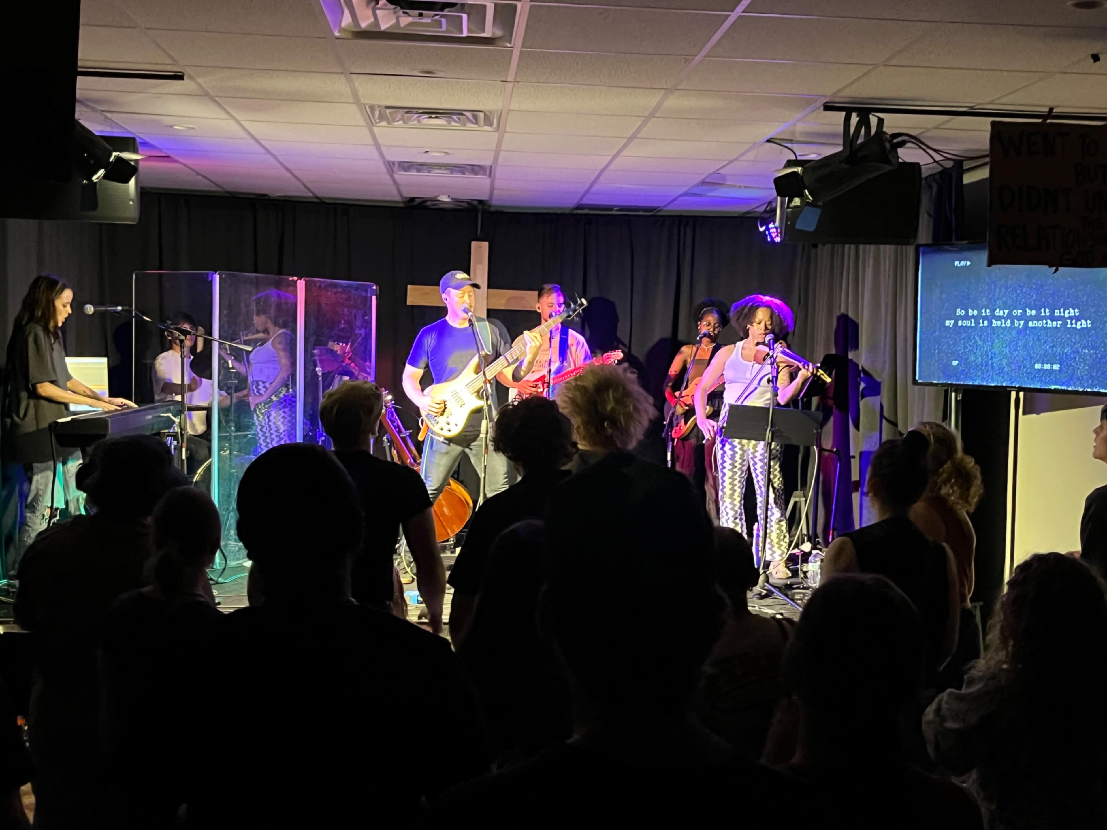 A band performs on stage with instruments including a keyboard, guitar, and violin, while an audience watches in a dimly lit room.