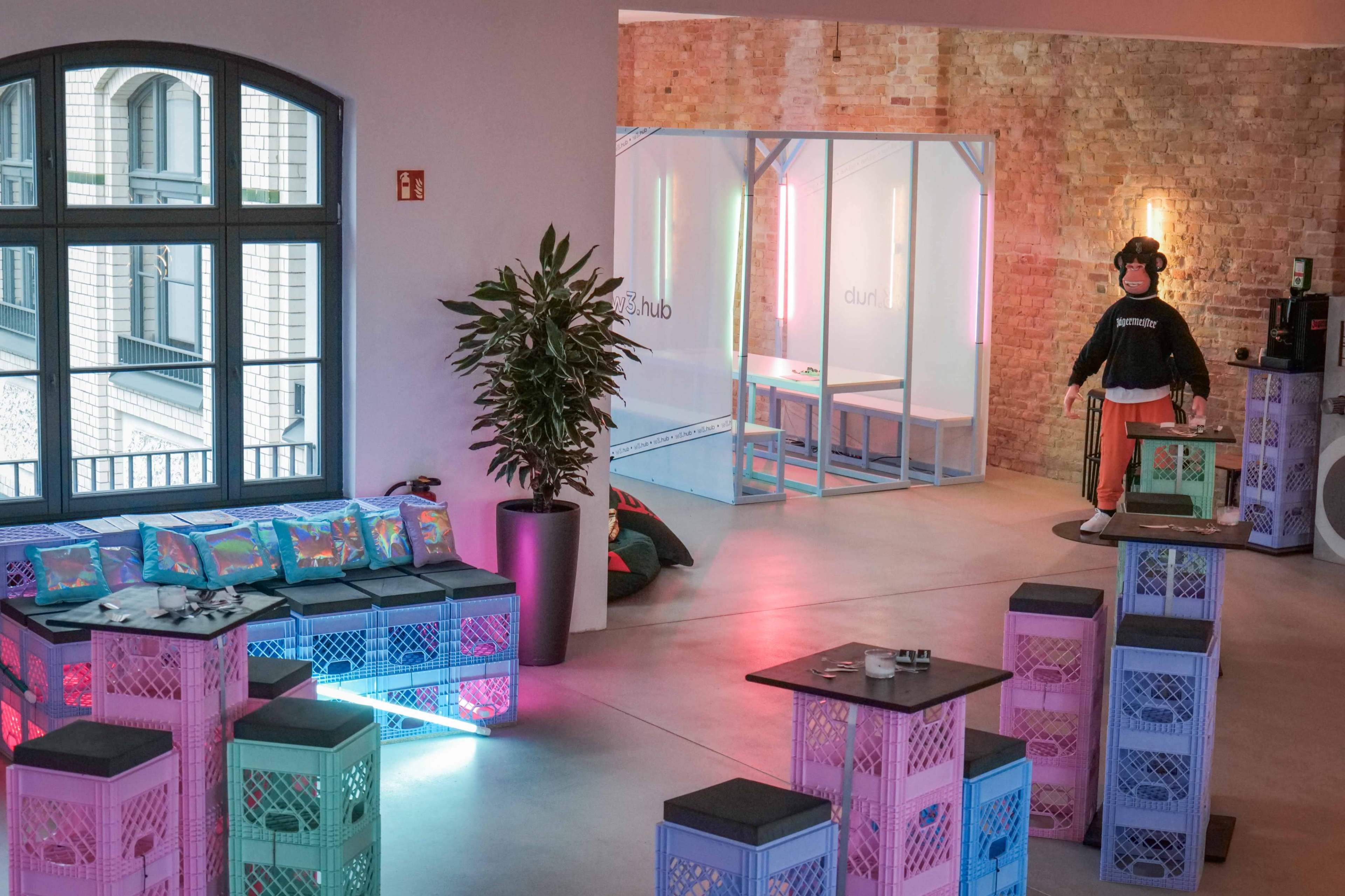 The image shows a modern indoor space featuring colorful crate furniture, a potted plant, and a clear partition separating areas for activities or meetings.