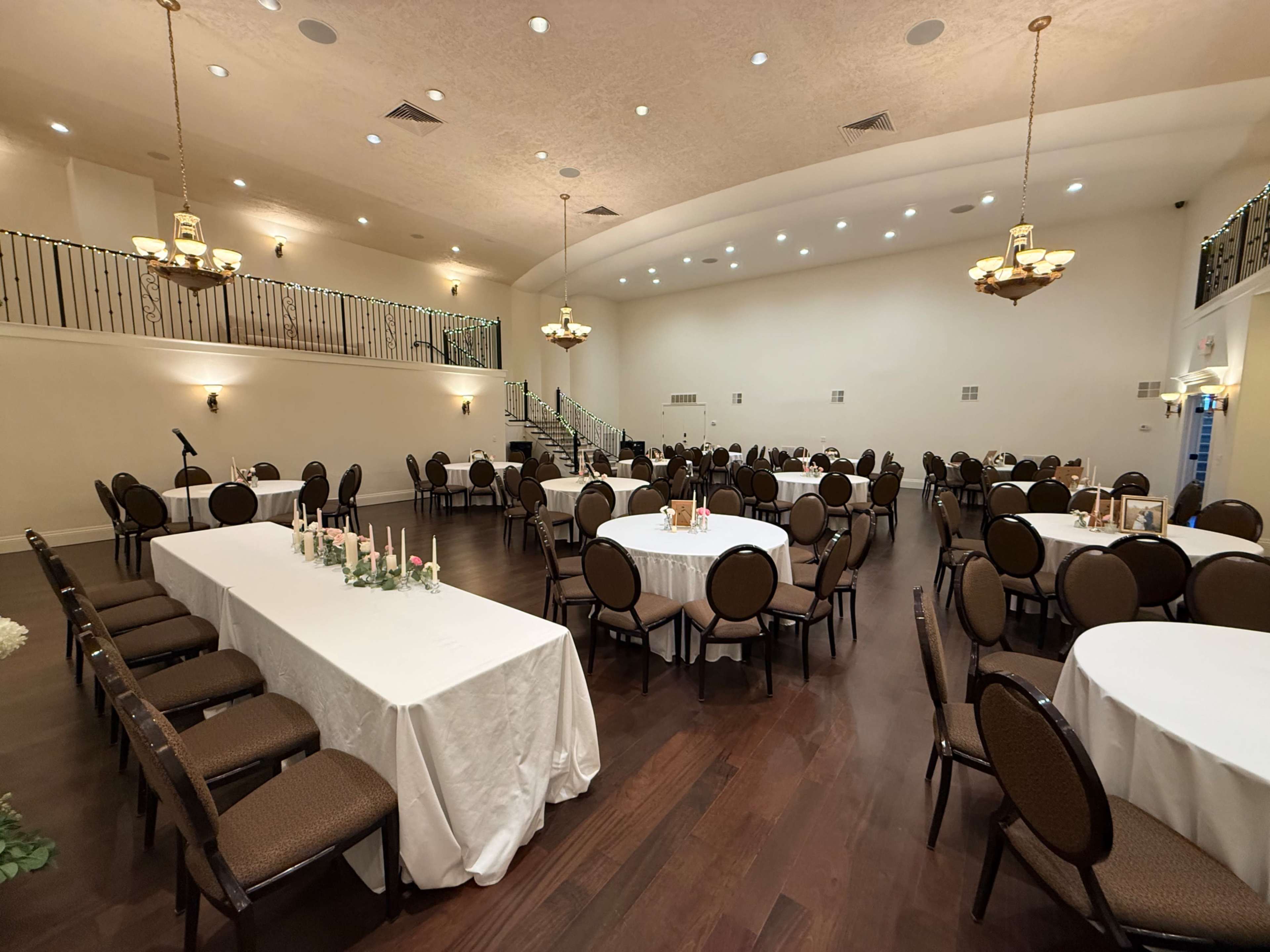 The image shows a spacious banquet hall with round tables set for dining and a long table arranged along one side, illuminated by several chandeliers.
