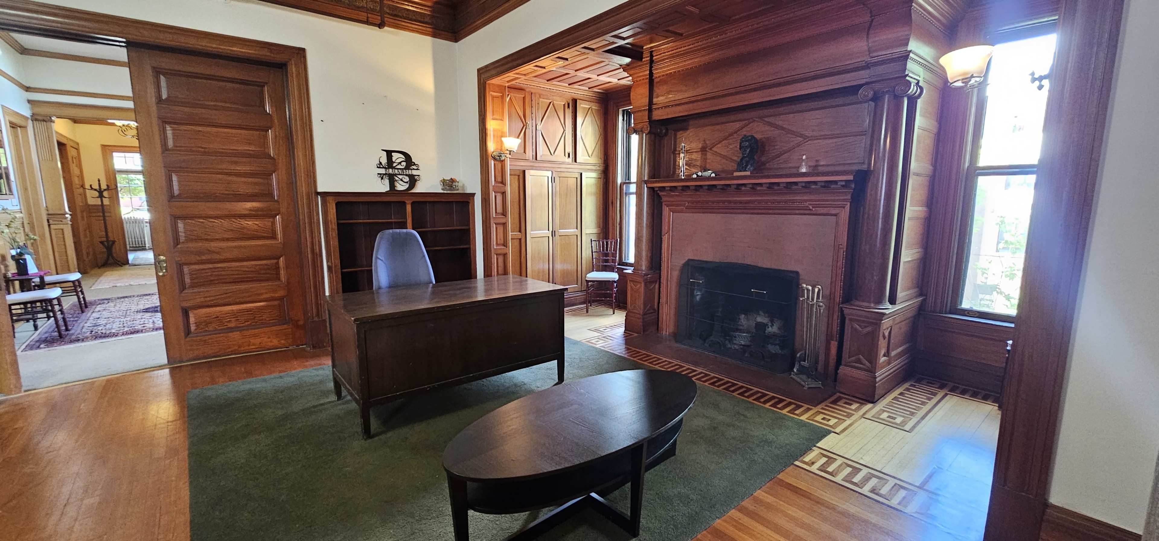 The image shows a room with a wooden desk and chair near a decorative fireplace, featuring intricate wooden paneling and a patterned floor.