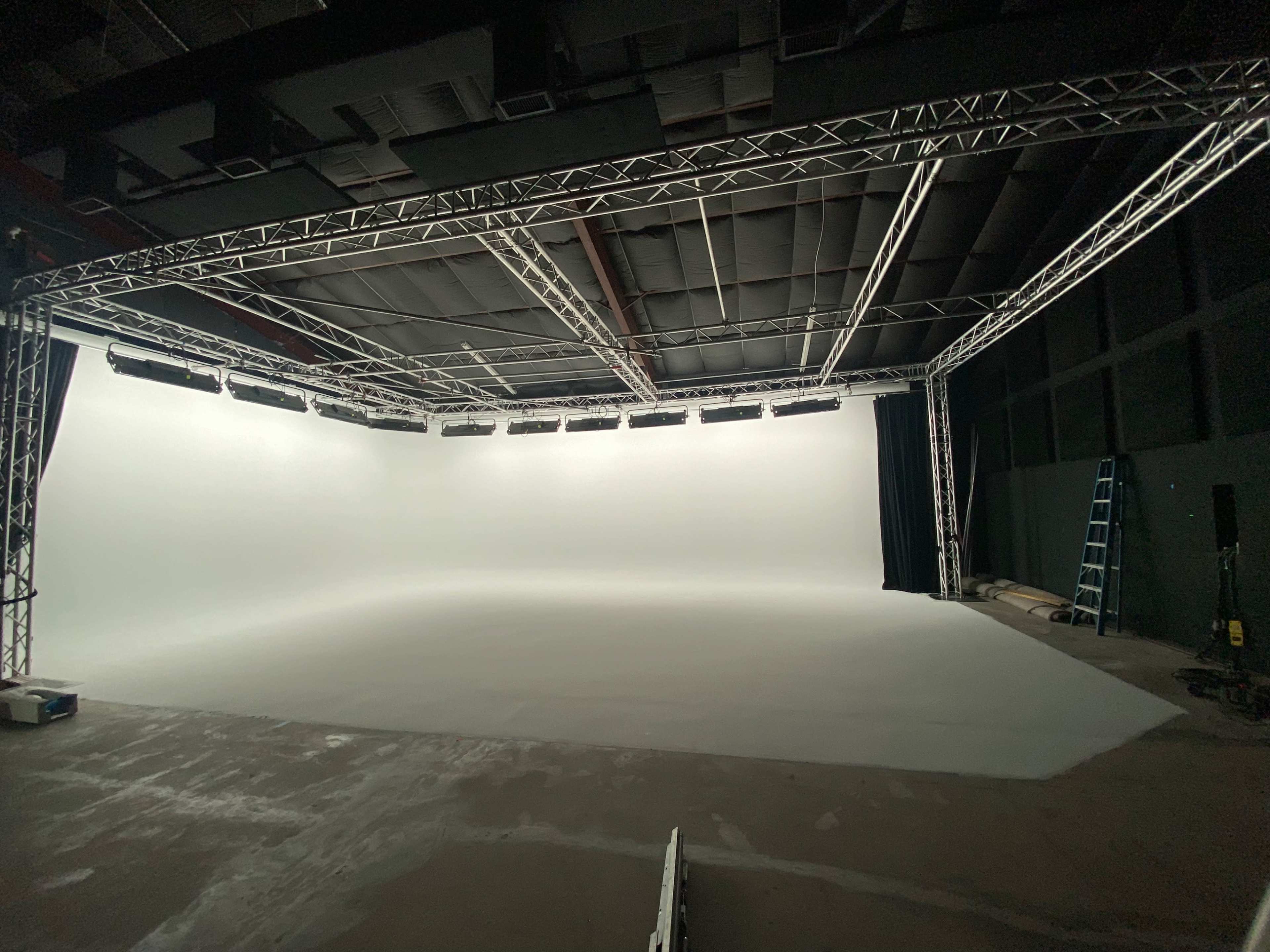 A spacious photography studio with a blank white backdrop, equipped with lighting fixtures and a ladder.