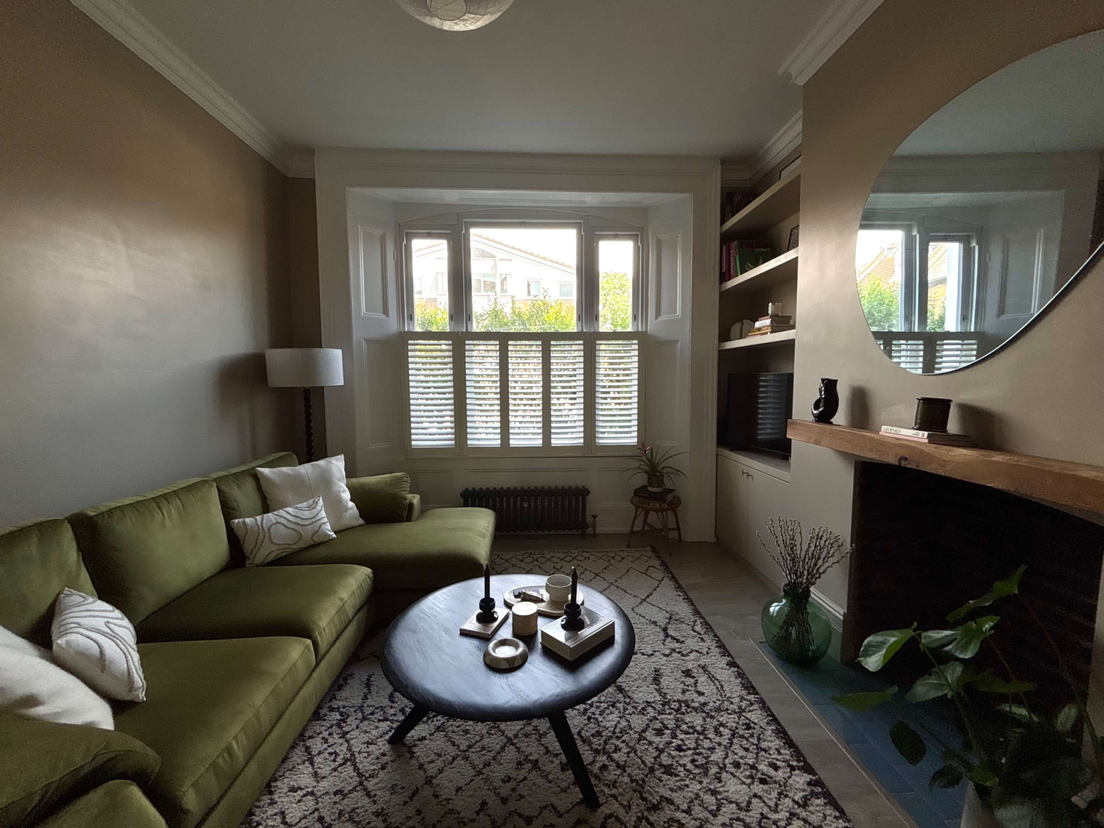 The image shows a cozy living room featuring a green sectional sofa, a round coffee table with decorative items, and a large window with shutters, complemented by a wall-mounted mirror and a bookshelf.