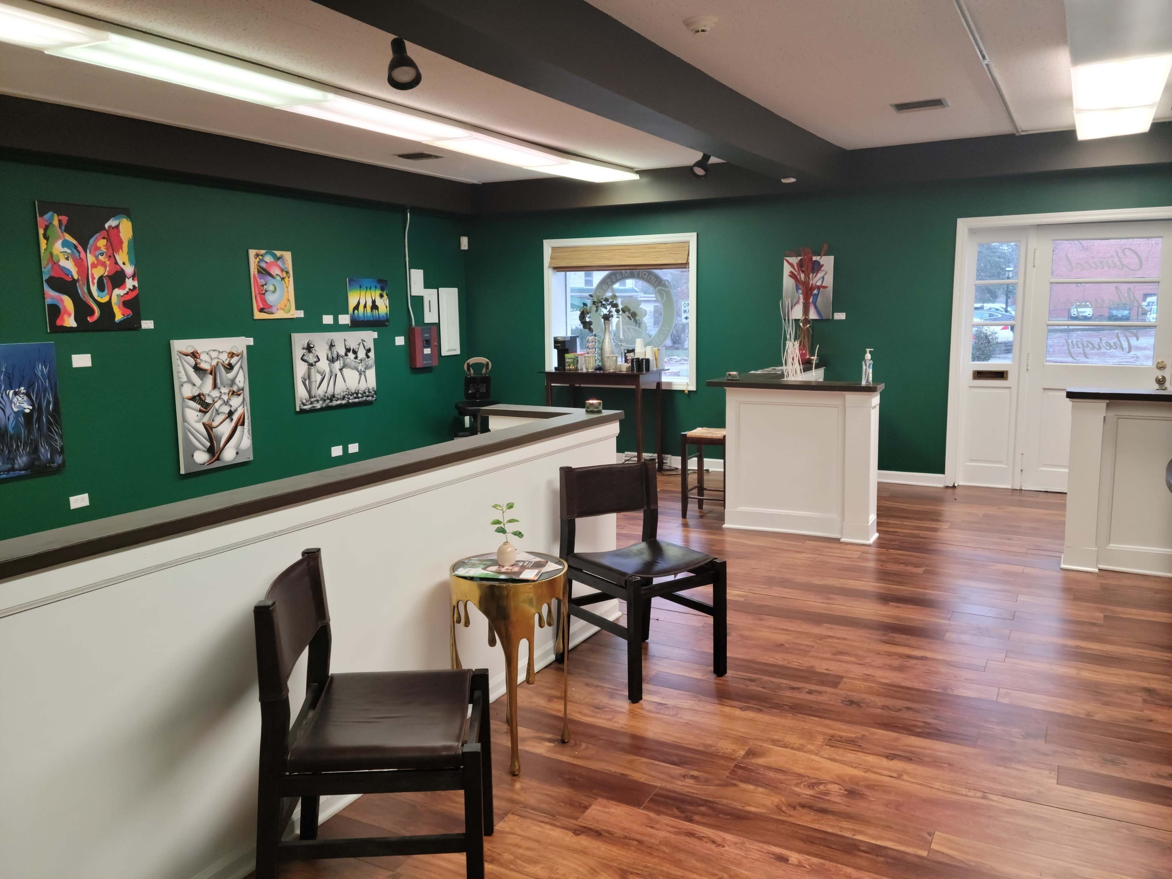 A brightly lit art gallery with green walls, featuring several paintings and a reception area with white counters and wooden chairs.