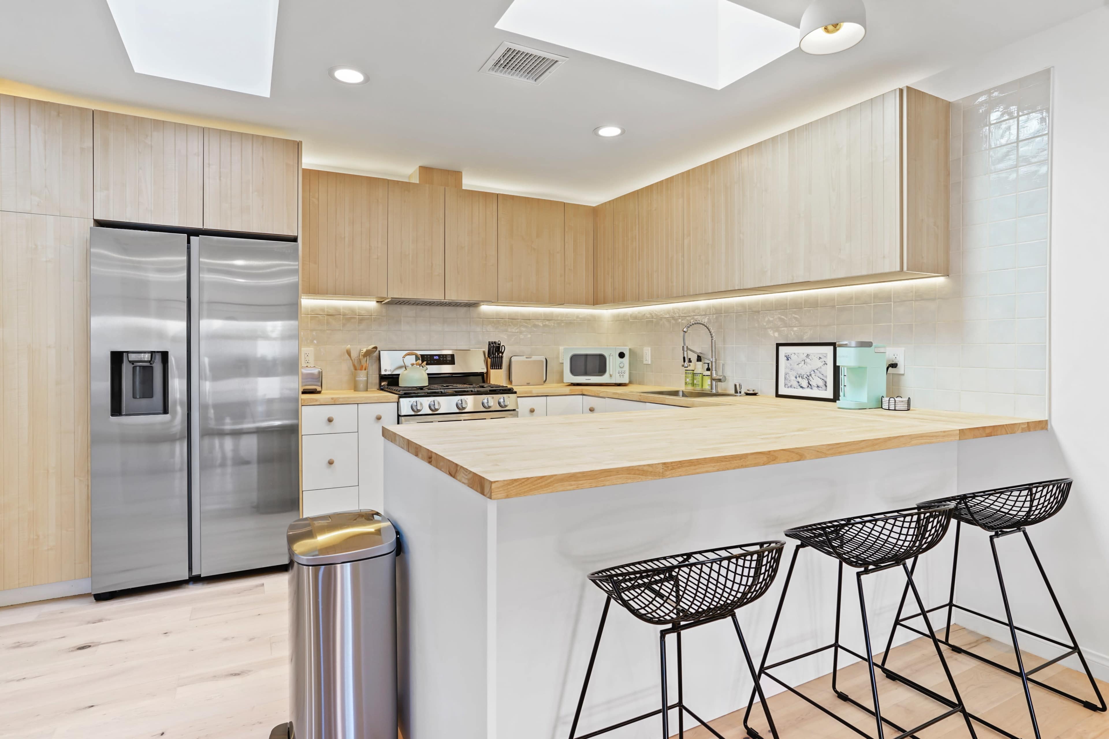 A modern kitchen featuring light wood cabinets, stainless steel appliances, a large wooden countertop, and three black wire bar stools.
