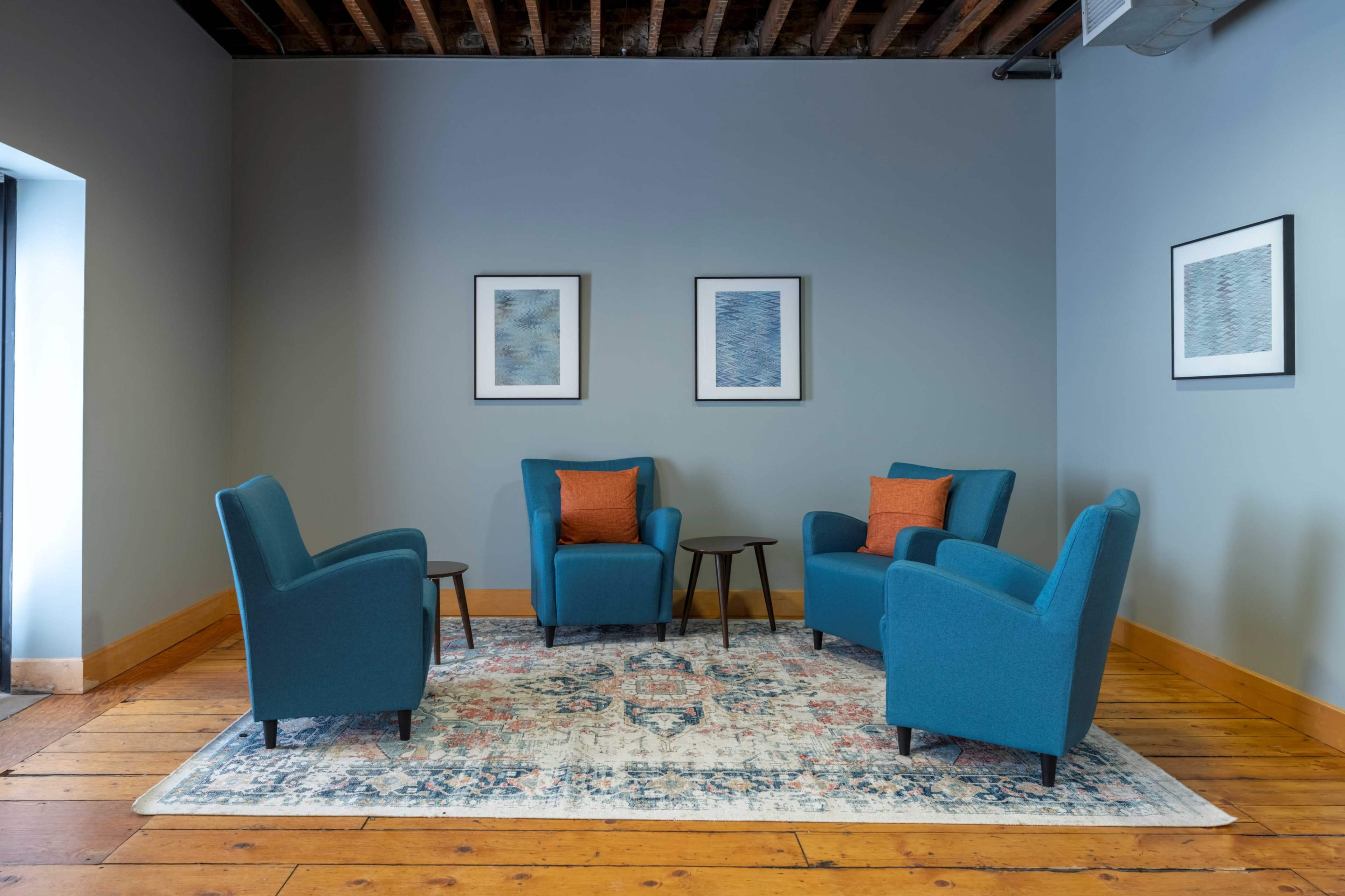 A seating area features four teal chairs arranged around two small tables on a patterned rug against a gray wall with framed artwork.