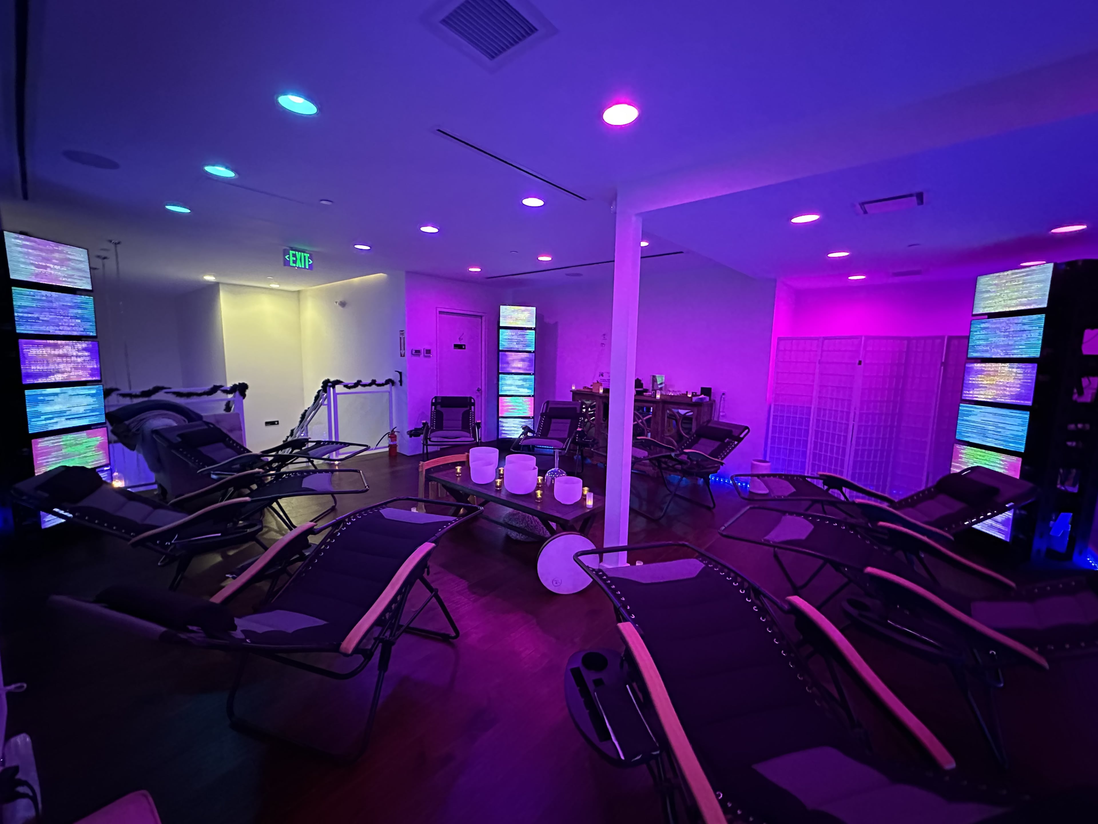 The image shows a dimly lit relaxation room with reclining lounge chairs and colorful illuminated screens lining the walls.